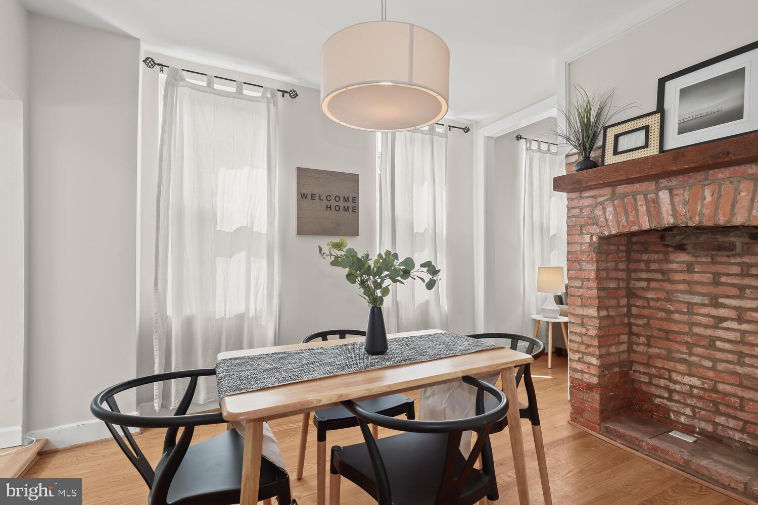 Staged dining room with a wood table, black chairs, sheer curtains, and a brick fireplace.