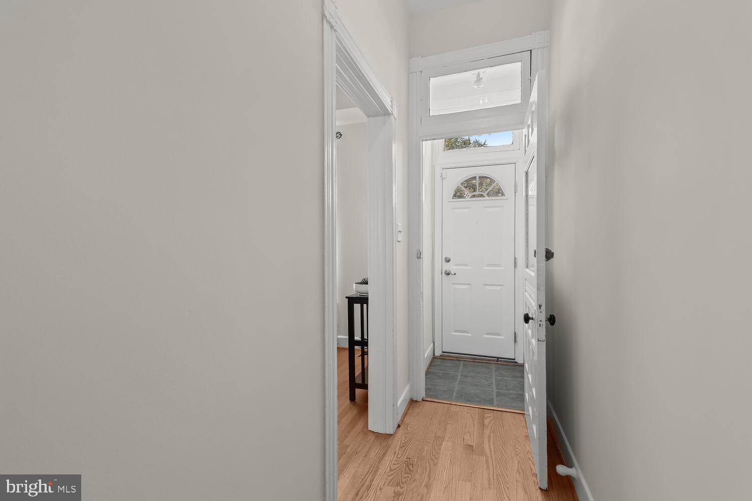 Entry hall with light wood floors, gray tile by the white front door, and a small table visible through a doorway.
