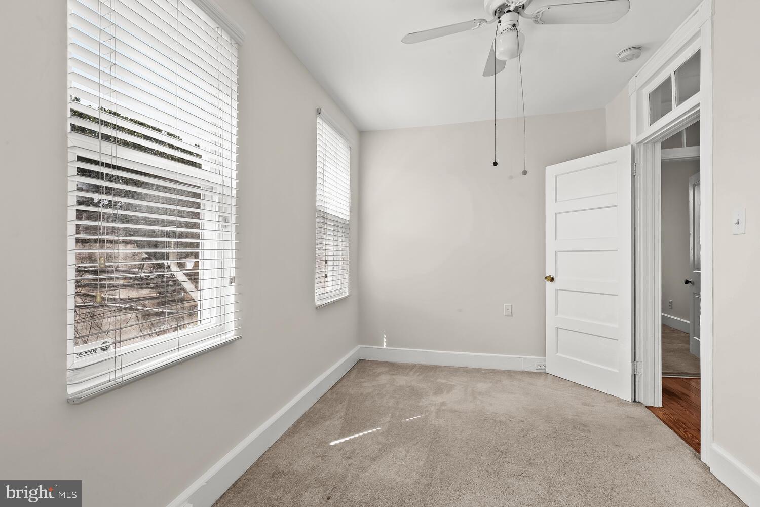 Empty carpeted bedroom with two blinded windows, a ceiling fan, and an open door to the hall.