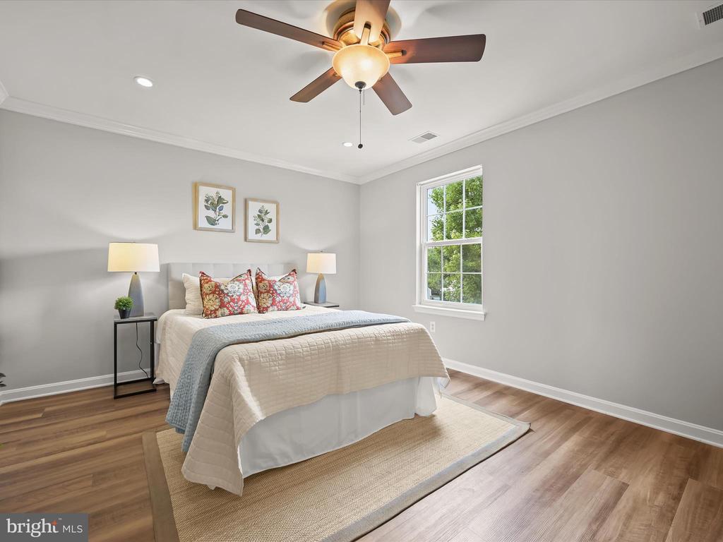 Staged bedroom with a cream-quilted bed, colorful floral pillows, ceiling fan, and hardwood floors.
