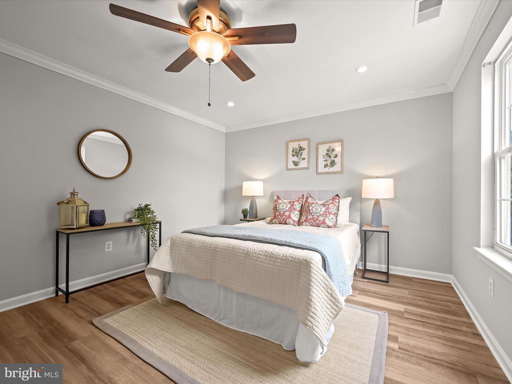 Staged bedroom with a white quilted bed, patterned pillows, ceiling fan, and large window.
