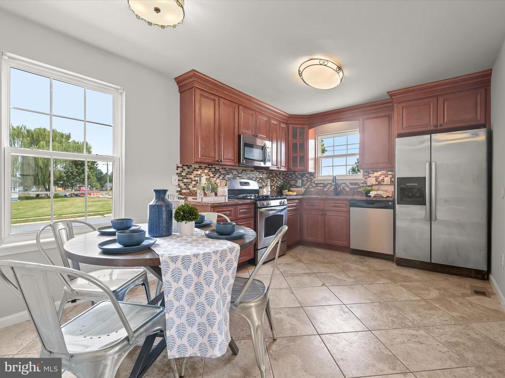 Staged kitchen with reddish-brown cabinets, stainless steel appliances, and a round dining table by large windows.