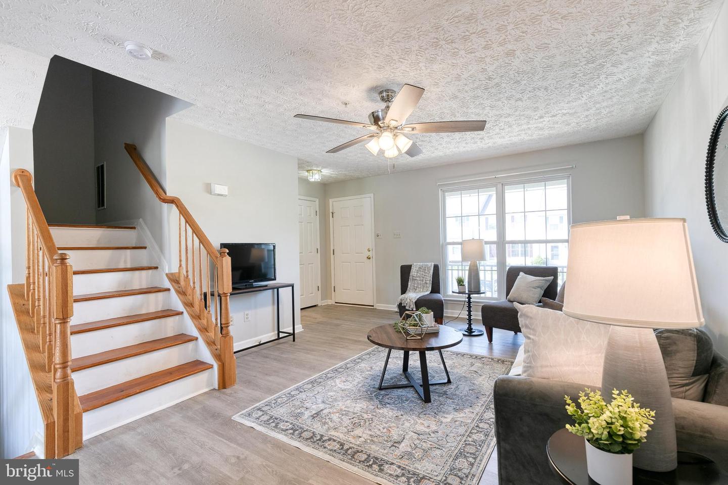Furnished living room with a wooden-railed staircase, gray seating, and large windows.