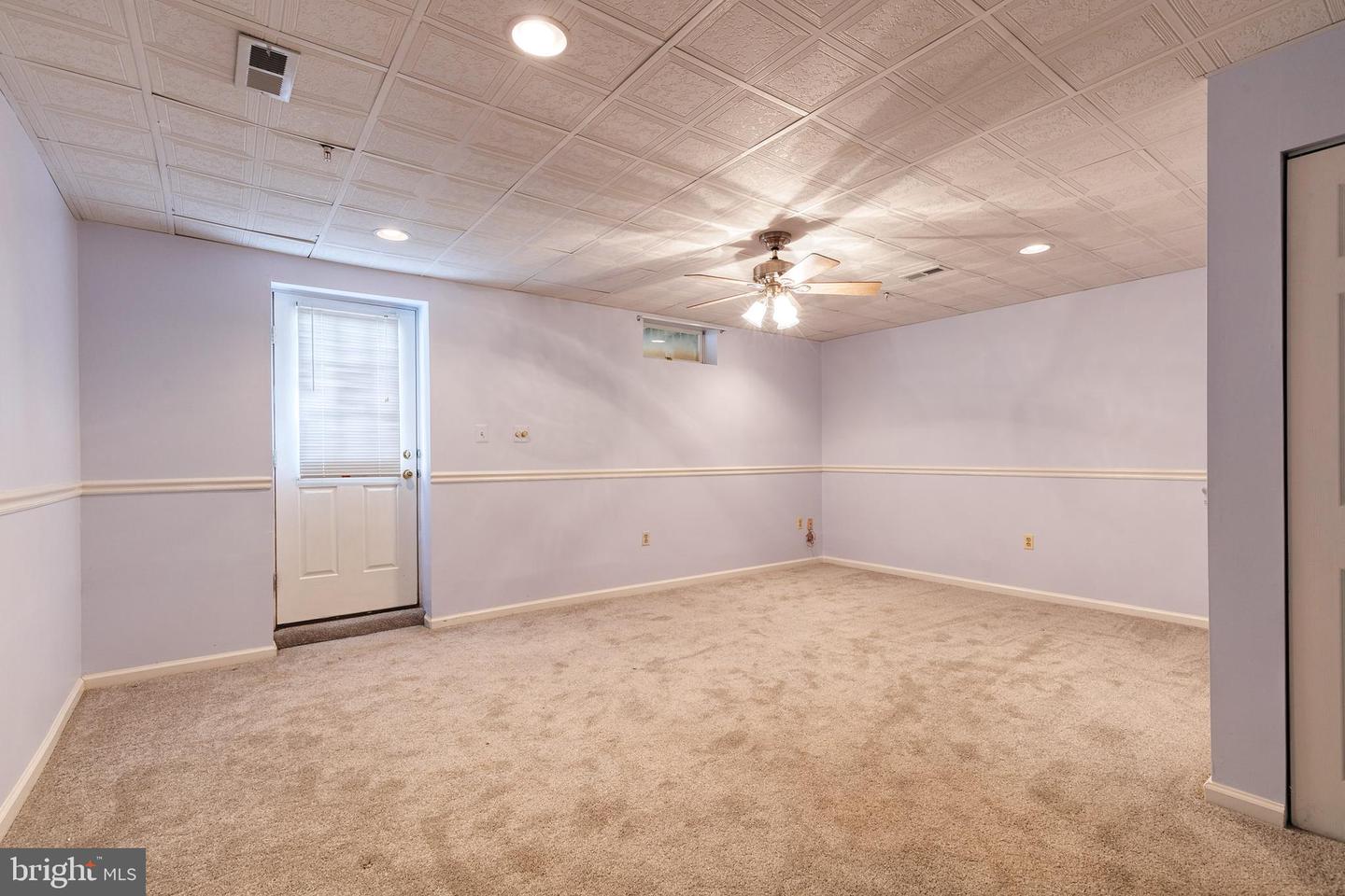 Empty carpeted room with a ceiling fan, patterned drop ceiling, and door with a window blind.