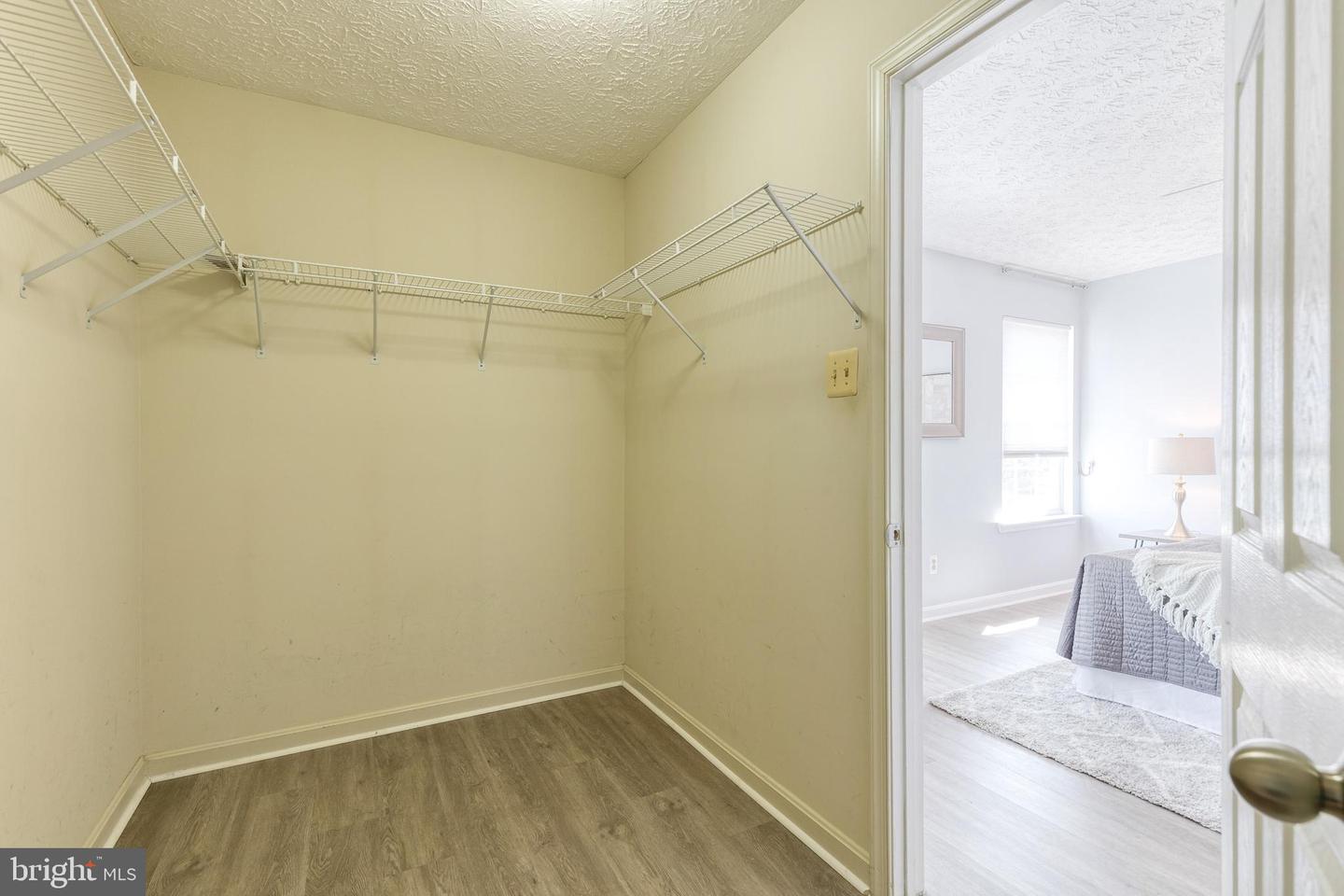 Walk-in closet with wire shelving and wood-look flooring, opening into a furnished bedroom.