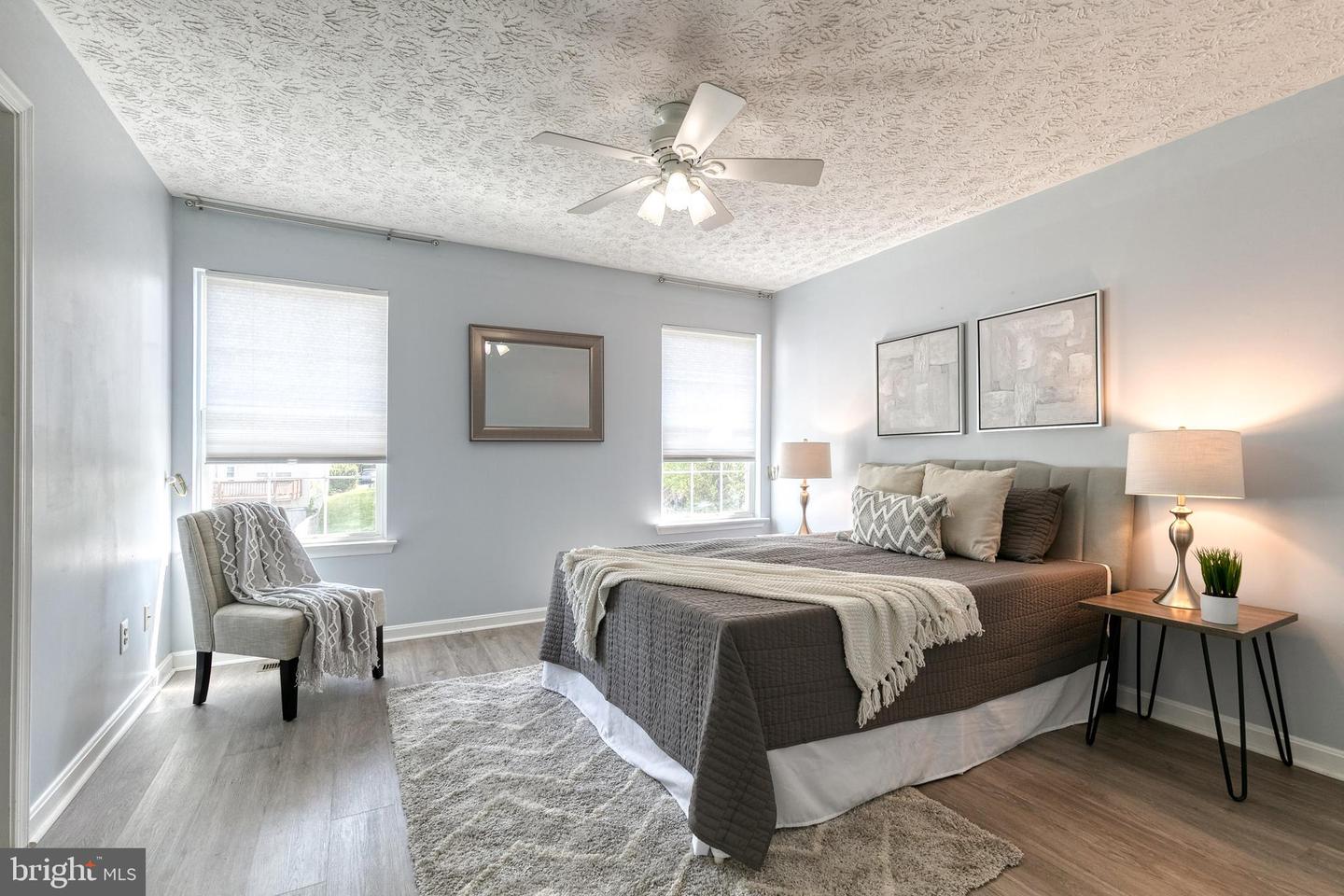 Staged bedroom with a gray upholstered bed, two windows, and a ceiling fan.