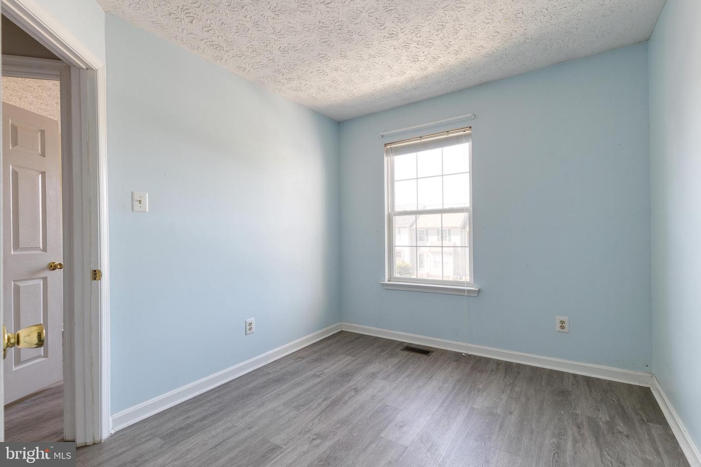 Empty bedroom with light blue walls, gray plank flooring, and a window overlooking a neighboring house.