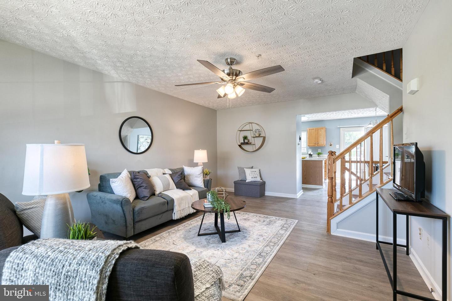 Staged living room with a gray sofa, patterned rug, and wooden staircase beside the kitchen.