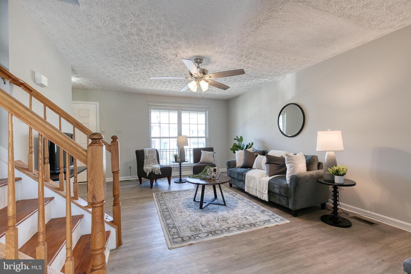 Staged living room with gray sofa, patterned rug, wooden staircase, and ceiling fan.