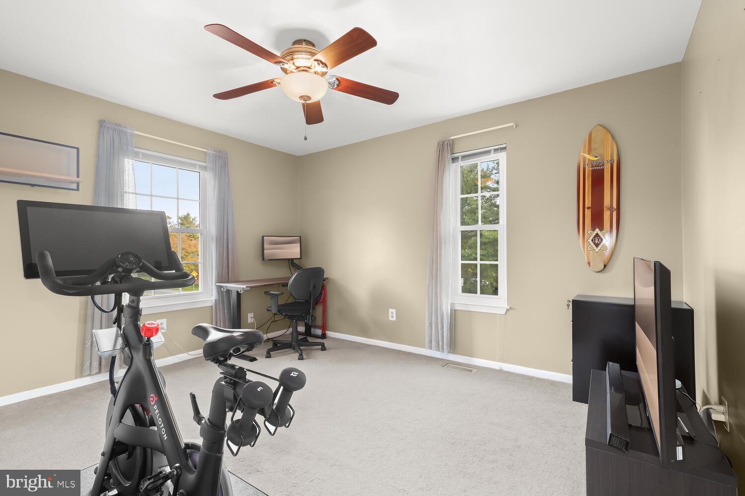 Home gym and workspace with a Peloton bike, adjustable weights, desk, and two windows.