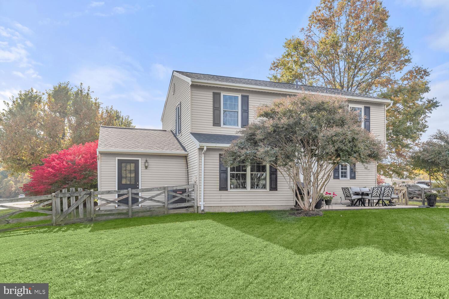 Two-story beige home with a fenced lawn and patio dining set beneath autumn trees.