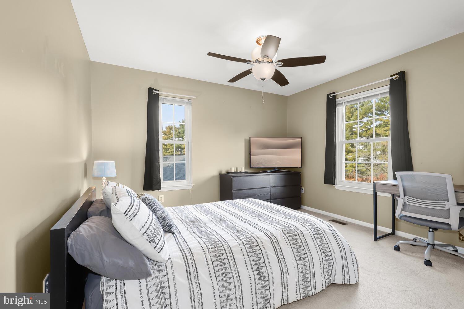 Bedroom with a patterned white bed, dark dresser and TV, ceiling fan, and windows overlooking trees.