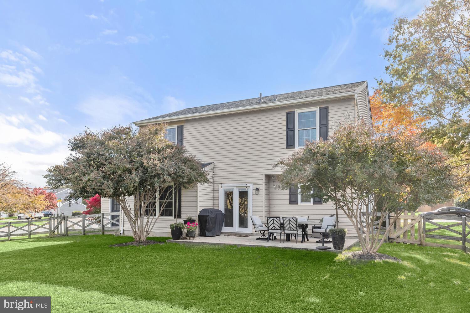 Two-story beige house with a patio dining set, grill, fenced yard, and autumn trees.