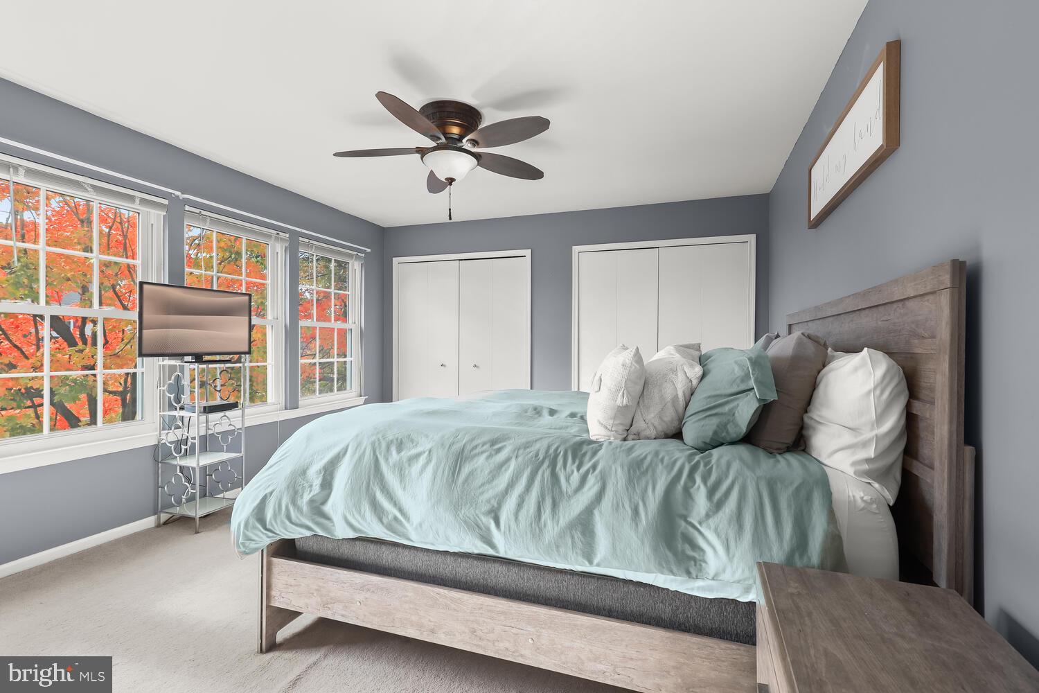 Staged bedroom with pale aqua bedding, gray walls, and windows overlooking autumn foliage.