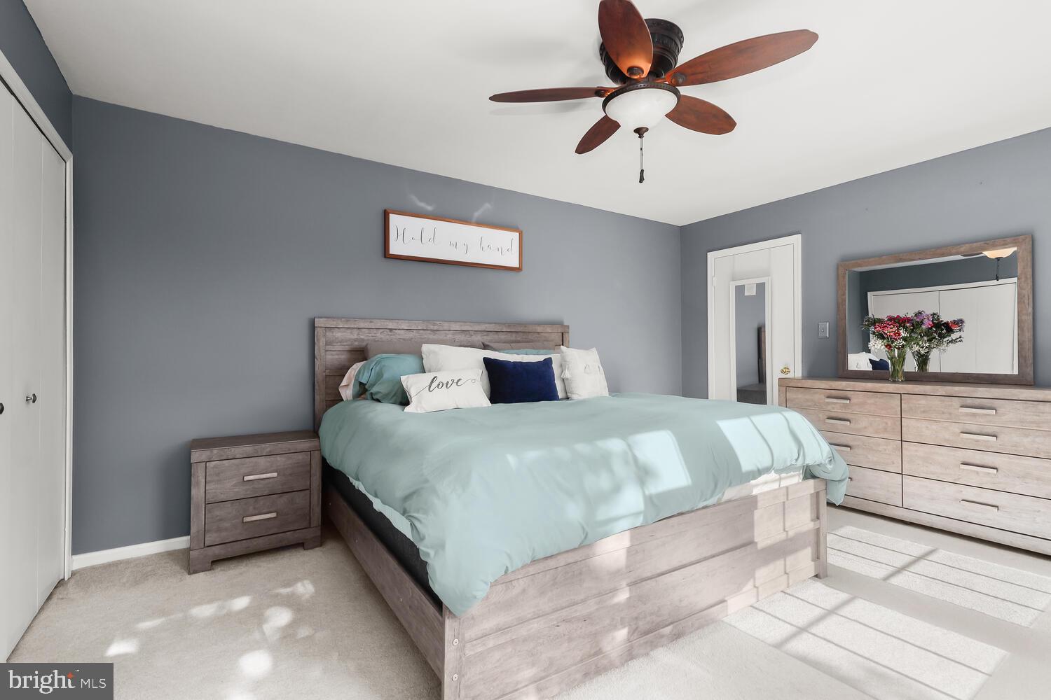 Staged bedroom with pale aqua bedding, gray walls, light wood furniture, and sunlight on the carpet.