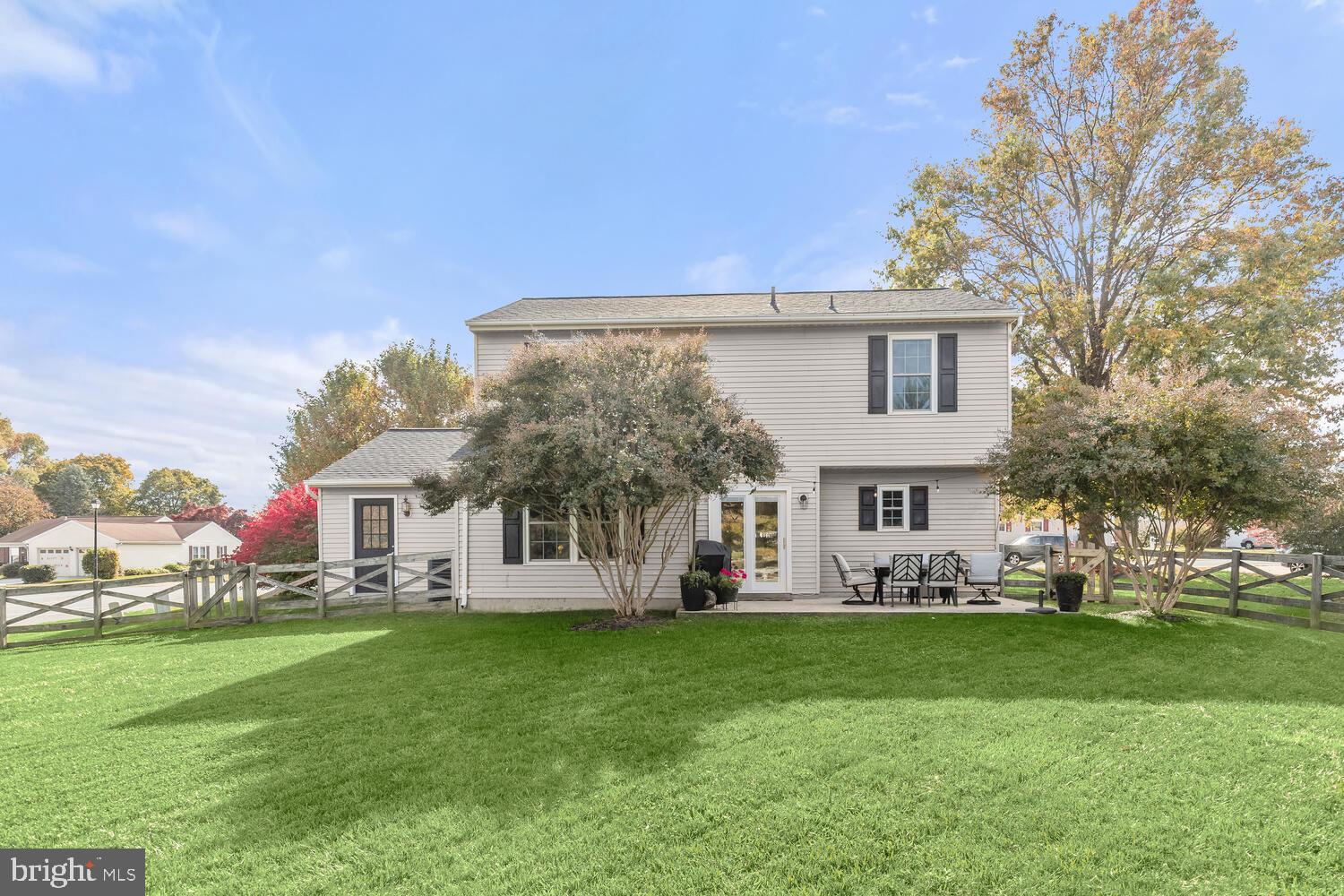 Rear of a two-story home with a fenced lawn and furnished patio beneath autumn trees.