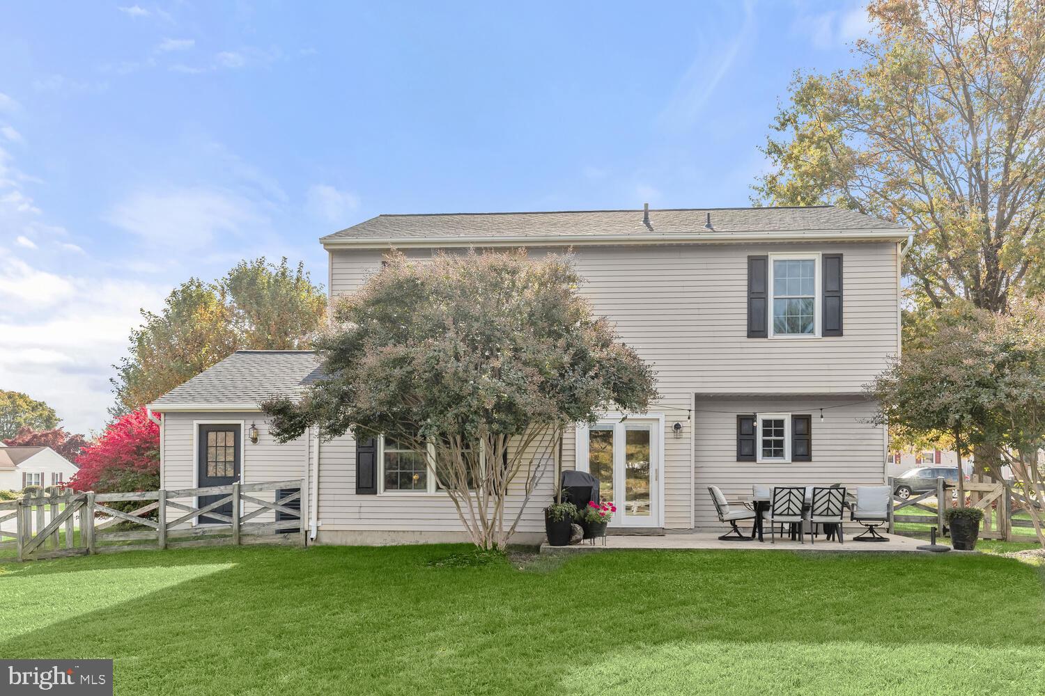 Rear of a beige two-story home with a fenced lawn and patio seating beneath autumn trees.