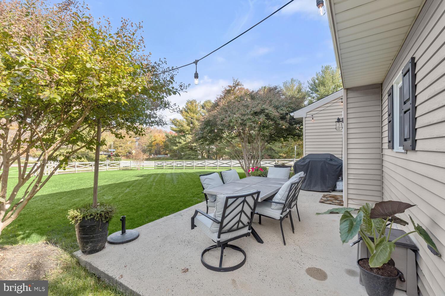 Furnished patio with outdoor dining set and covered grill beside a fenced lawn.