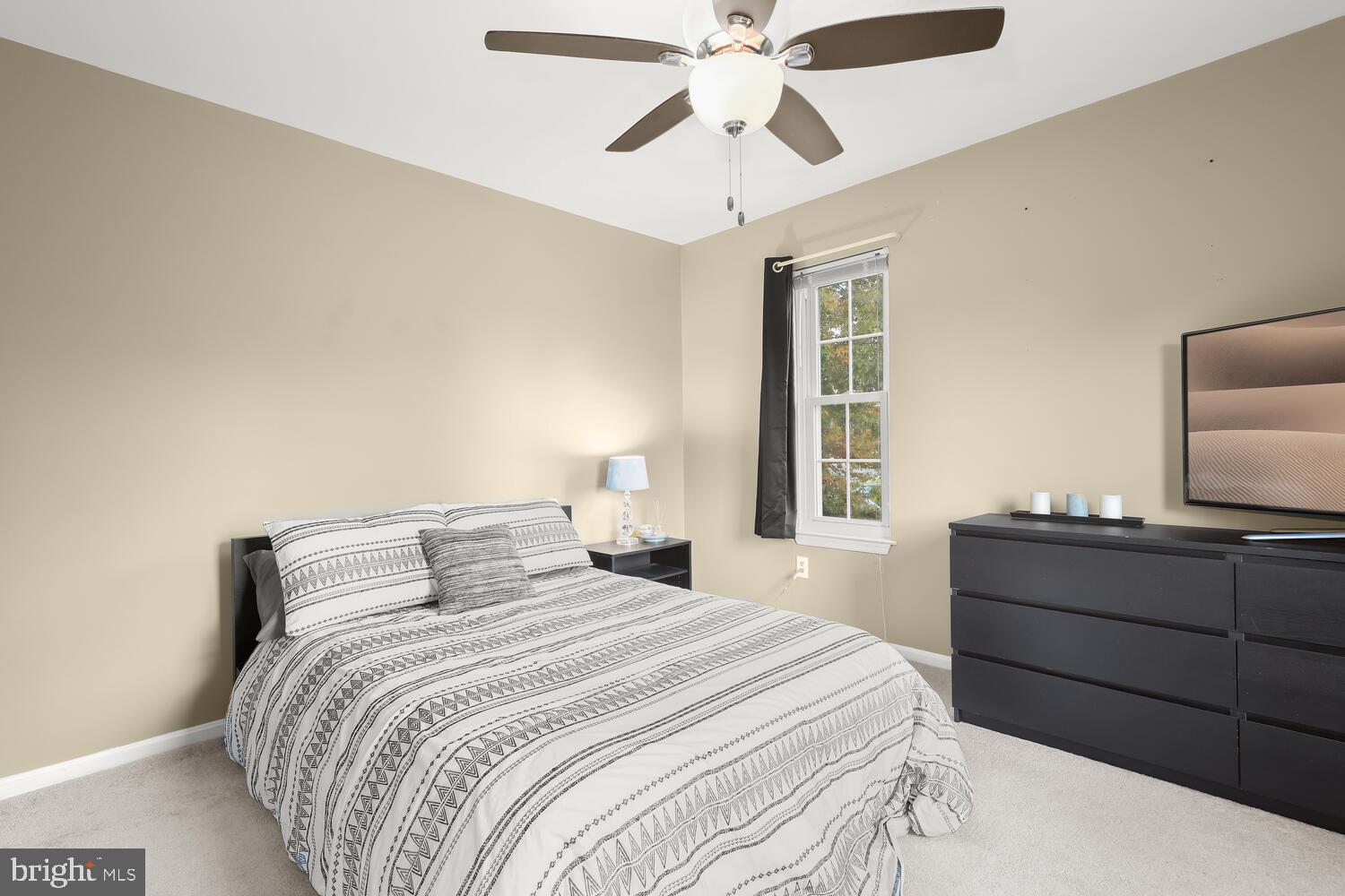 Furnished bedroom with a patterned bedspread, ceiling fan, window, and dark dresser with television.