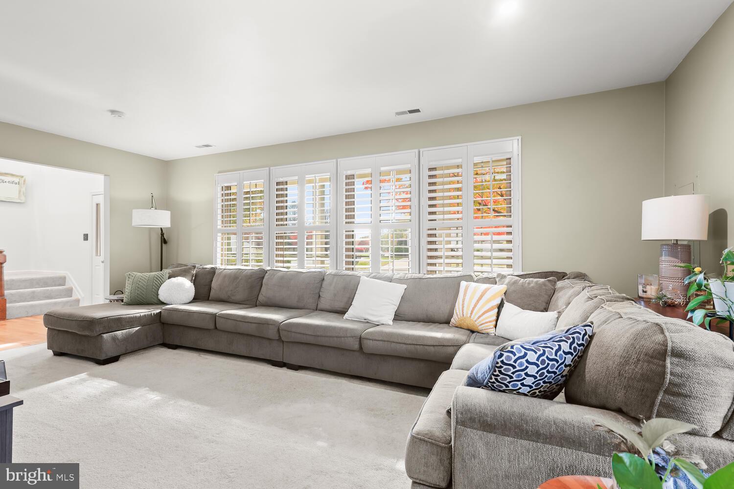 Living room with a gray sectional sofa, white plantation shutters, and autumn foliage outside.