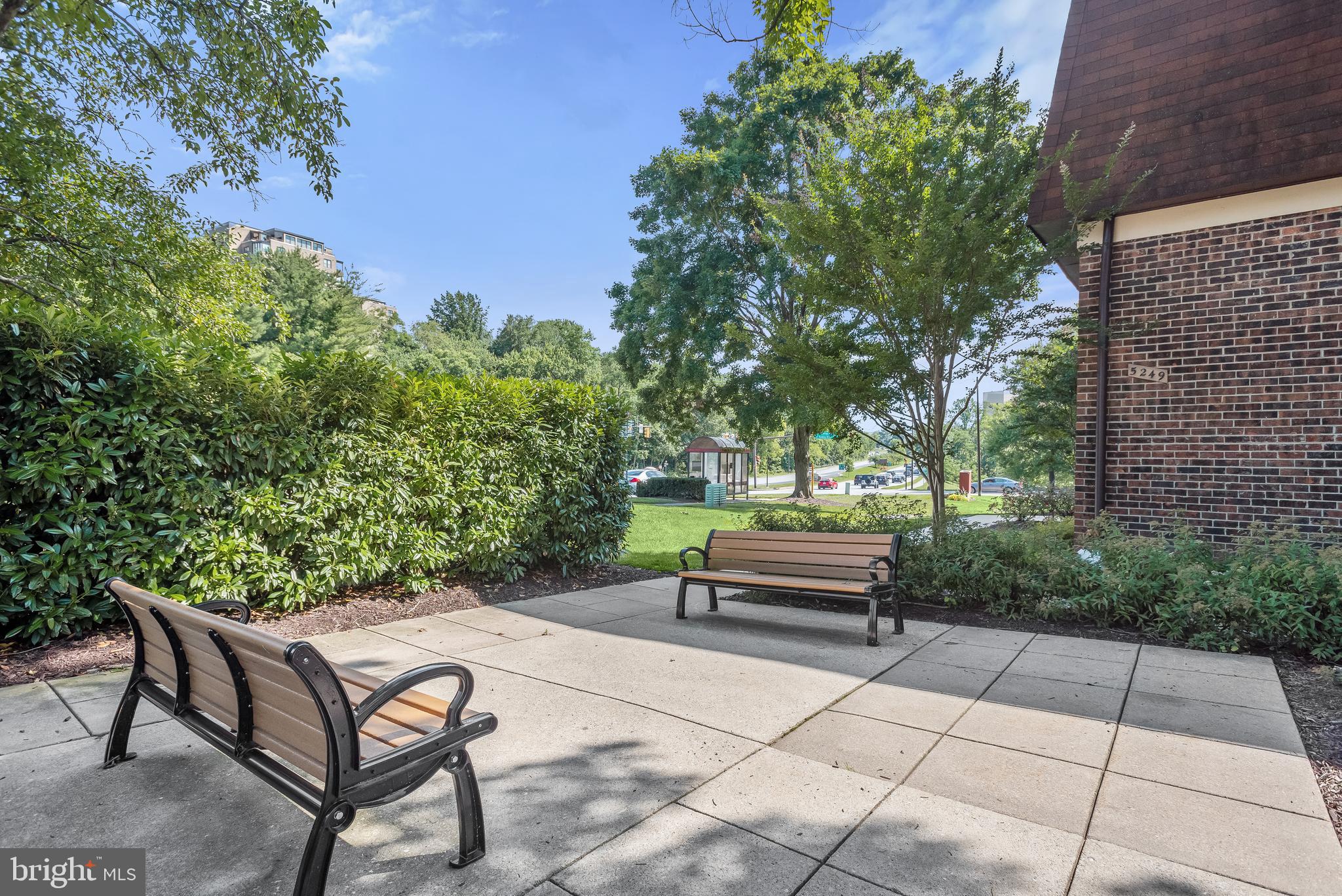 Outdoor seating area beside a brick building, with two benches and landscaped greenery.
