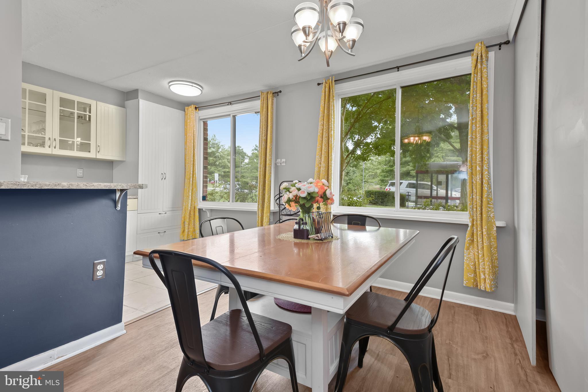Dining area with a wood-top table, black chairs, white cabinets, and large windows with yellow curtains.