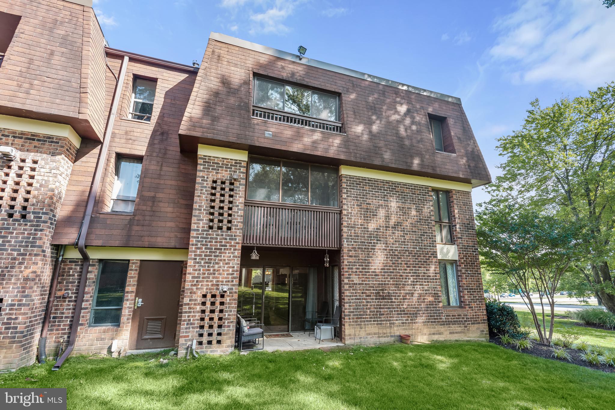 Condominium exterior with brick facade, ground-level patio, upper balcony, and mature trees.