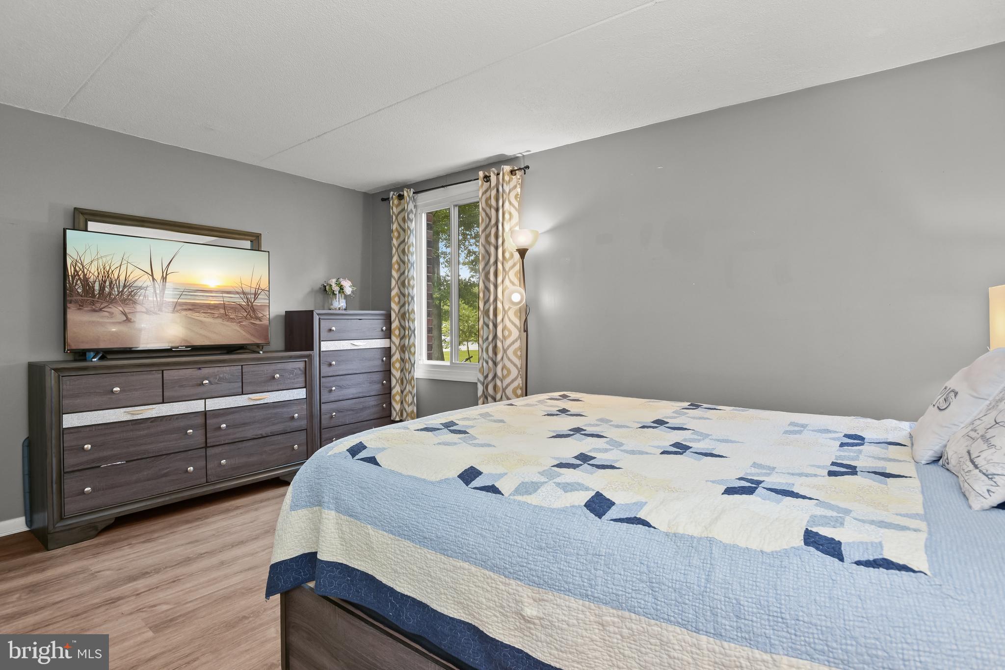 Bedroom with a blue-and-white quilted bed, dark wood dressers, and a window with patterned curtains.