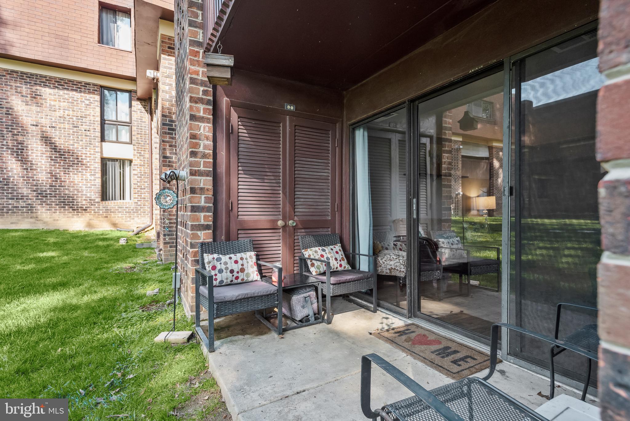 Ground-level apartment patio with wicker chairs, louvered doors, sliding glass doors, and a view of the lawn.