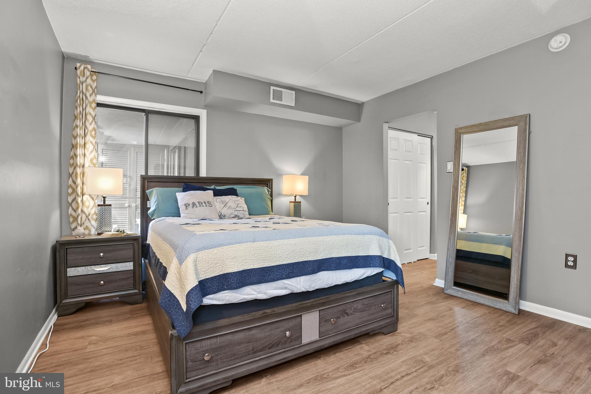 Furnished bedroom with a dark wood storage bed, blue-and-white quilt, and full-length mirror.
