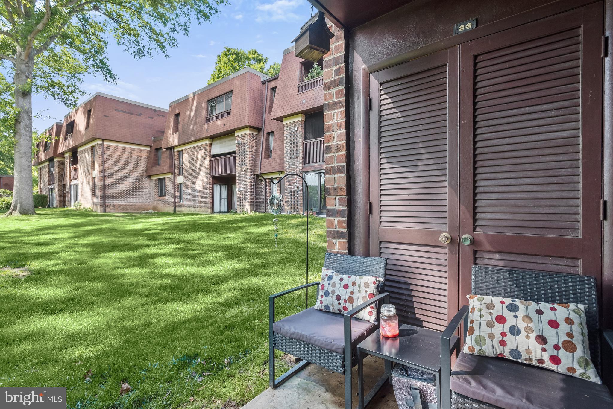 Brick condominium exterior beside a grassy courtyard, with a furnished patio and louvered doors.