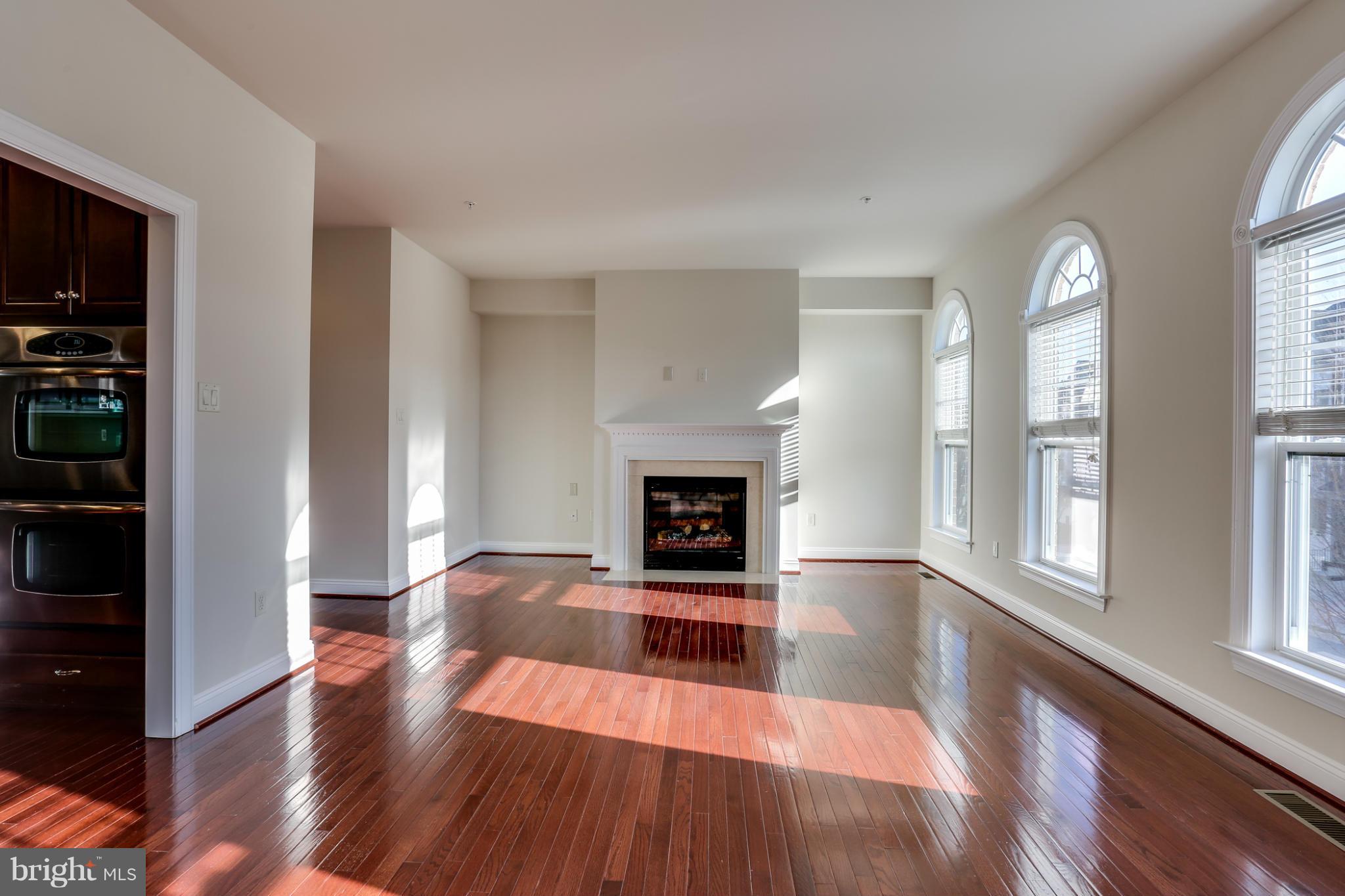 Empty living room with a fireplace, glossy hardwood floors, and tall arched windows.