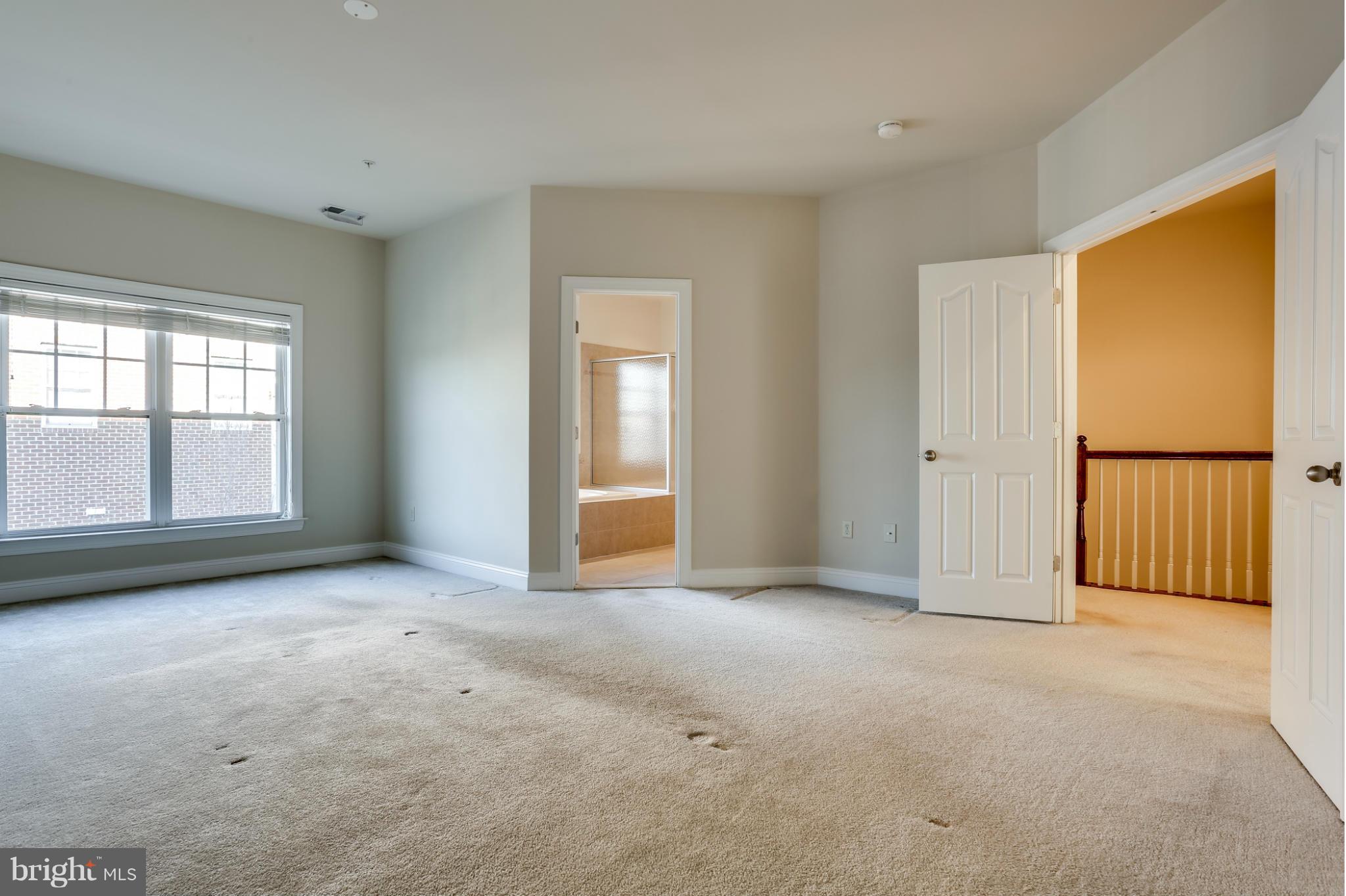Empty carpeted bedroom with a large window and open doorways to a bathroom and stair landing.