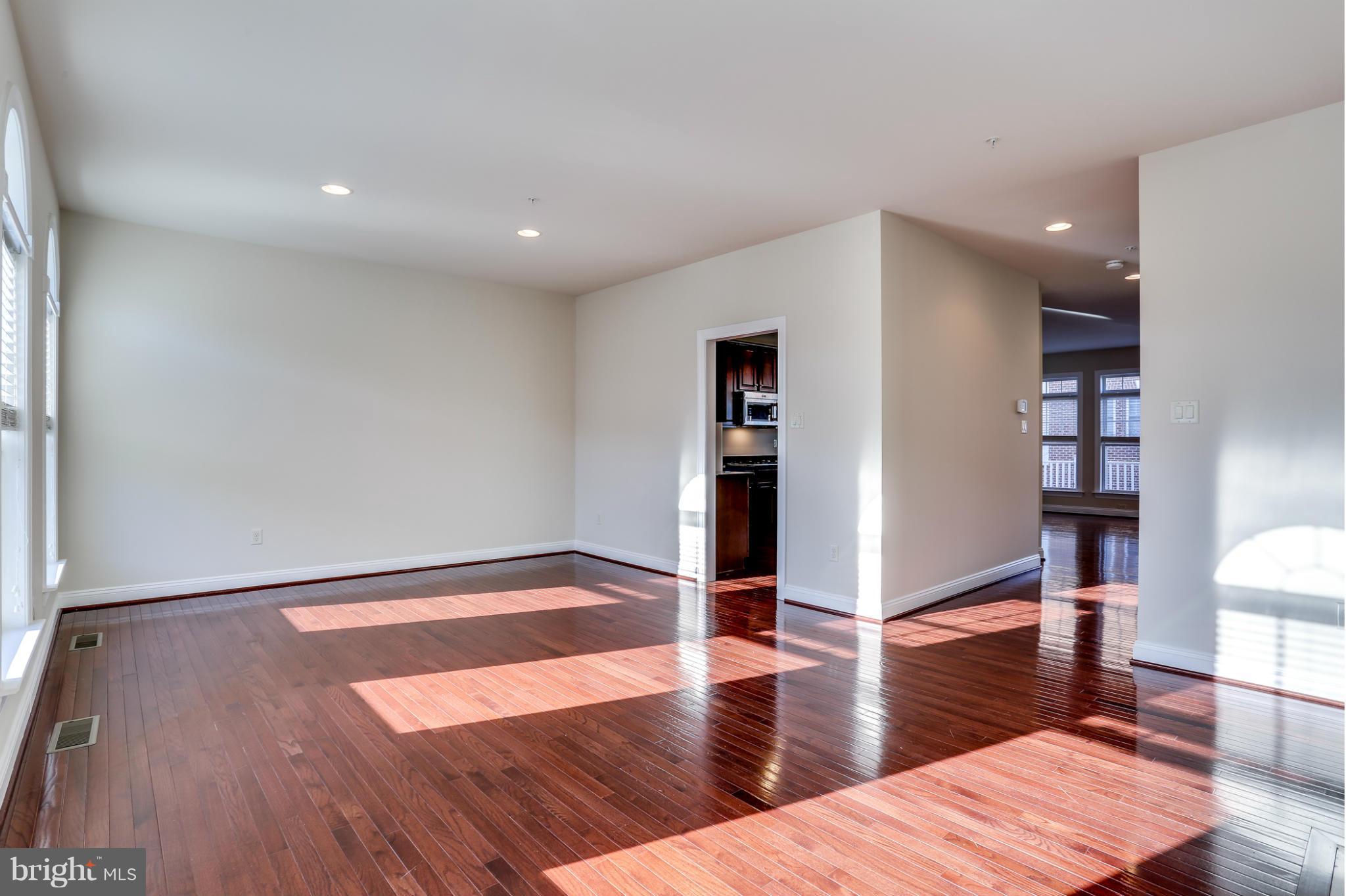 Vacant living room with glossy hardwood floors, tall windows, and sunlight streaming across the floor.