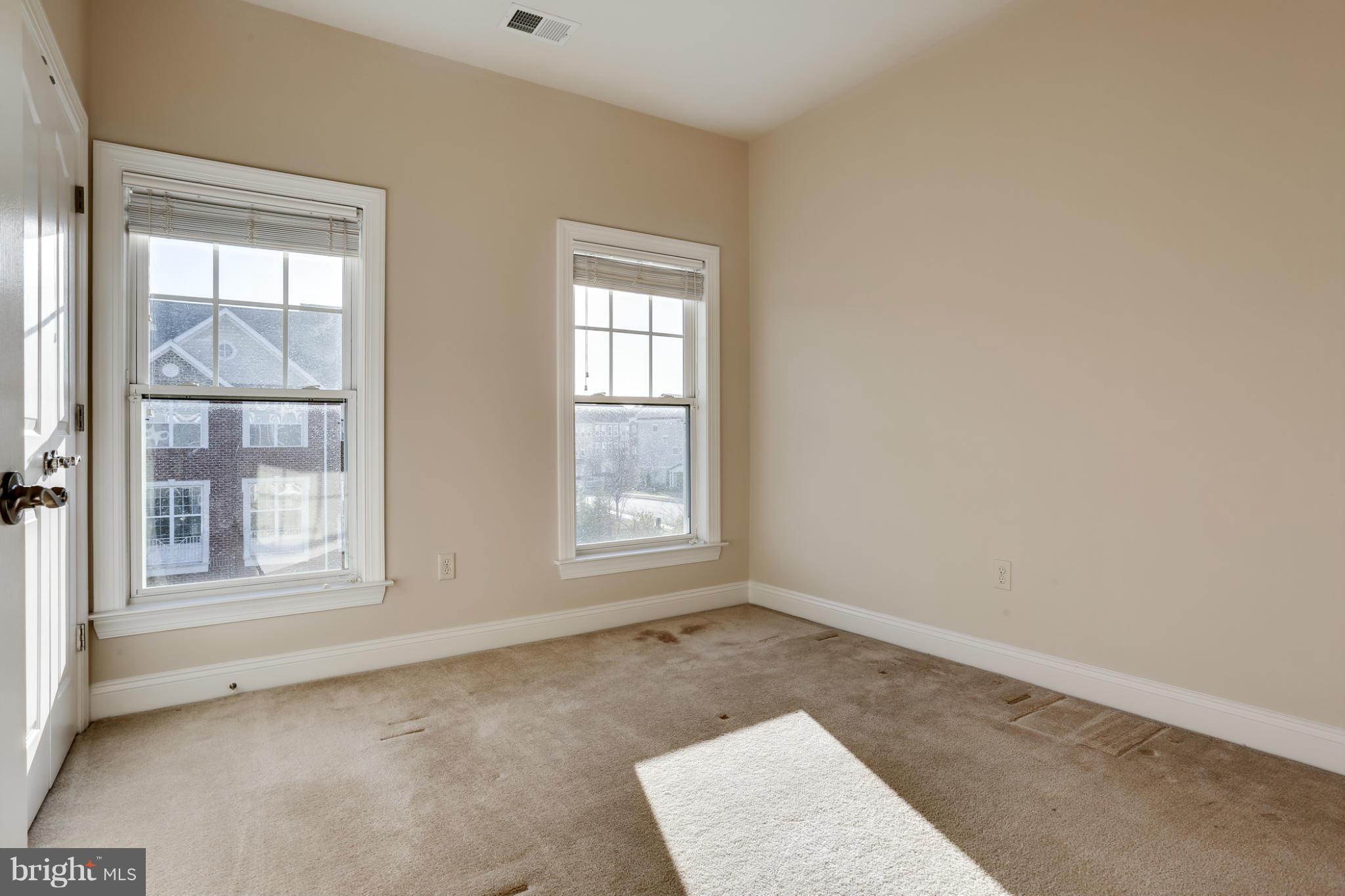 Empty carpeted room with two windows, blinds, and a bright patch of sunlight.