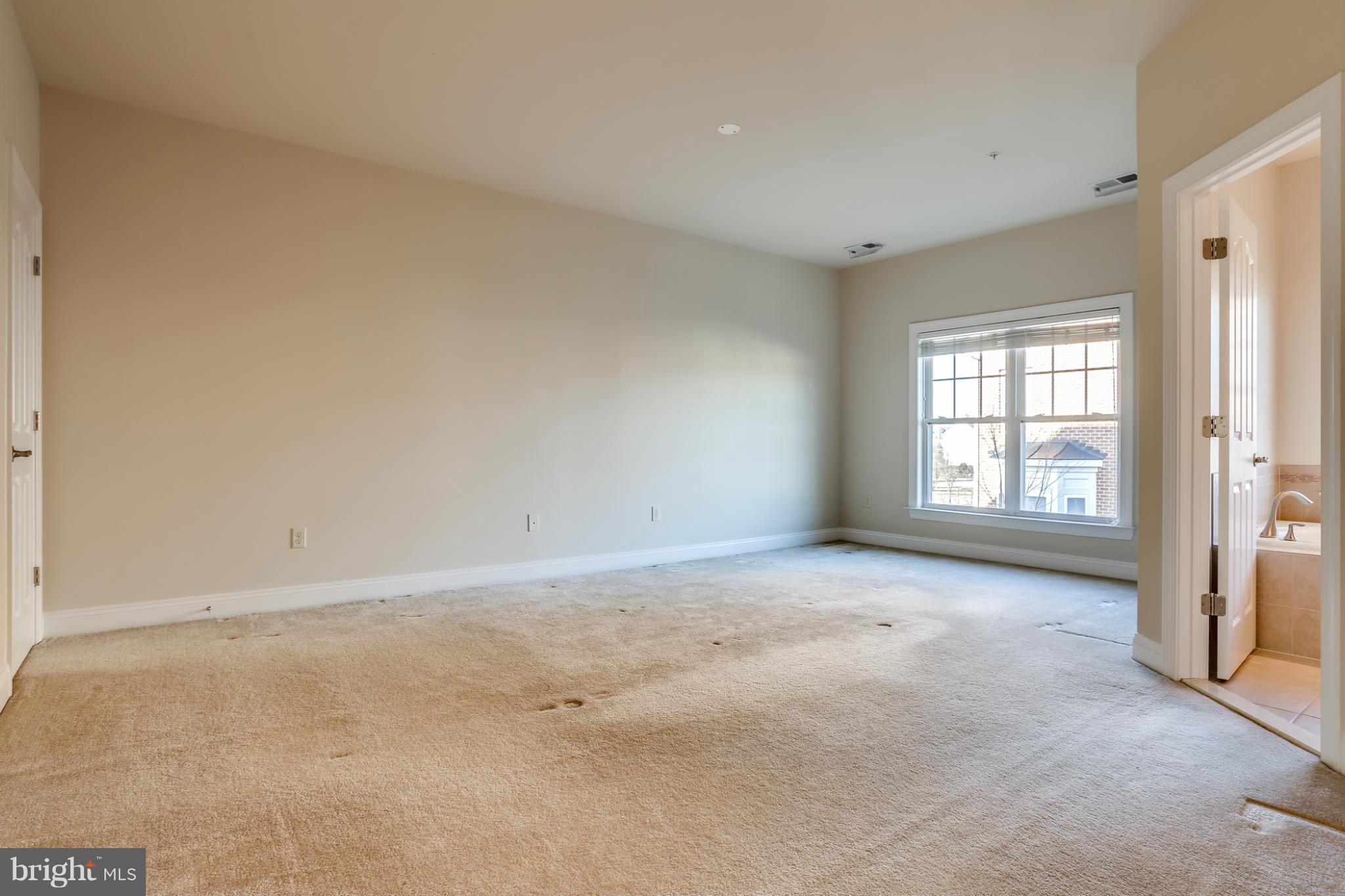 Empty carpeted bedroom with a large window and an open doorway to a tiled bathroom.