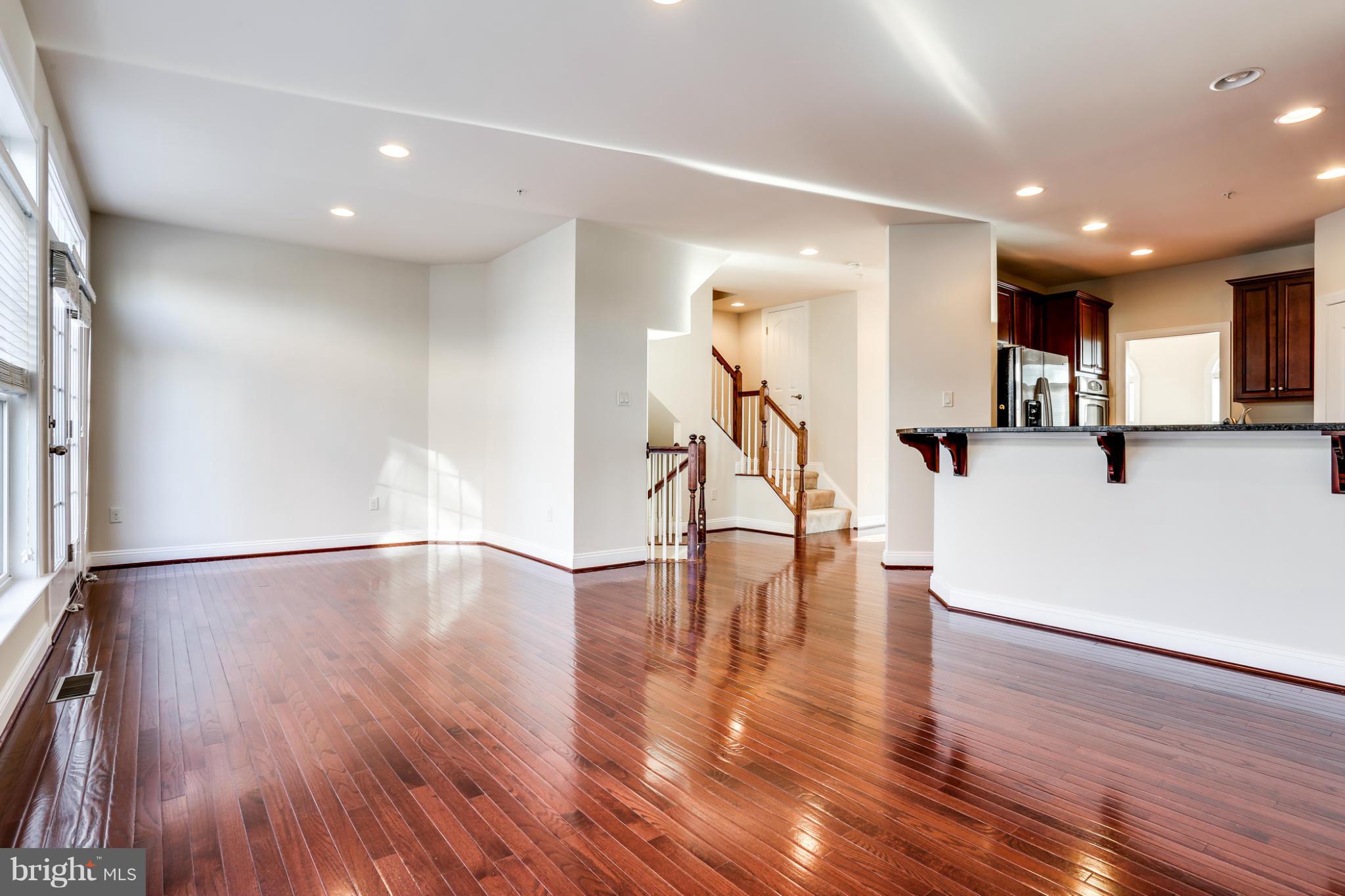 Open living and dining area with glossy hardwood floors, tall windows, staircase, and adjoining kitchen.