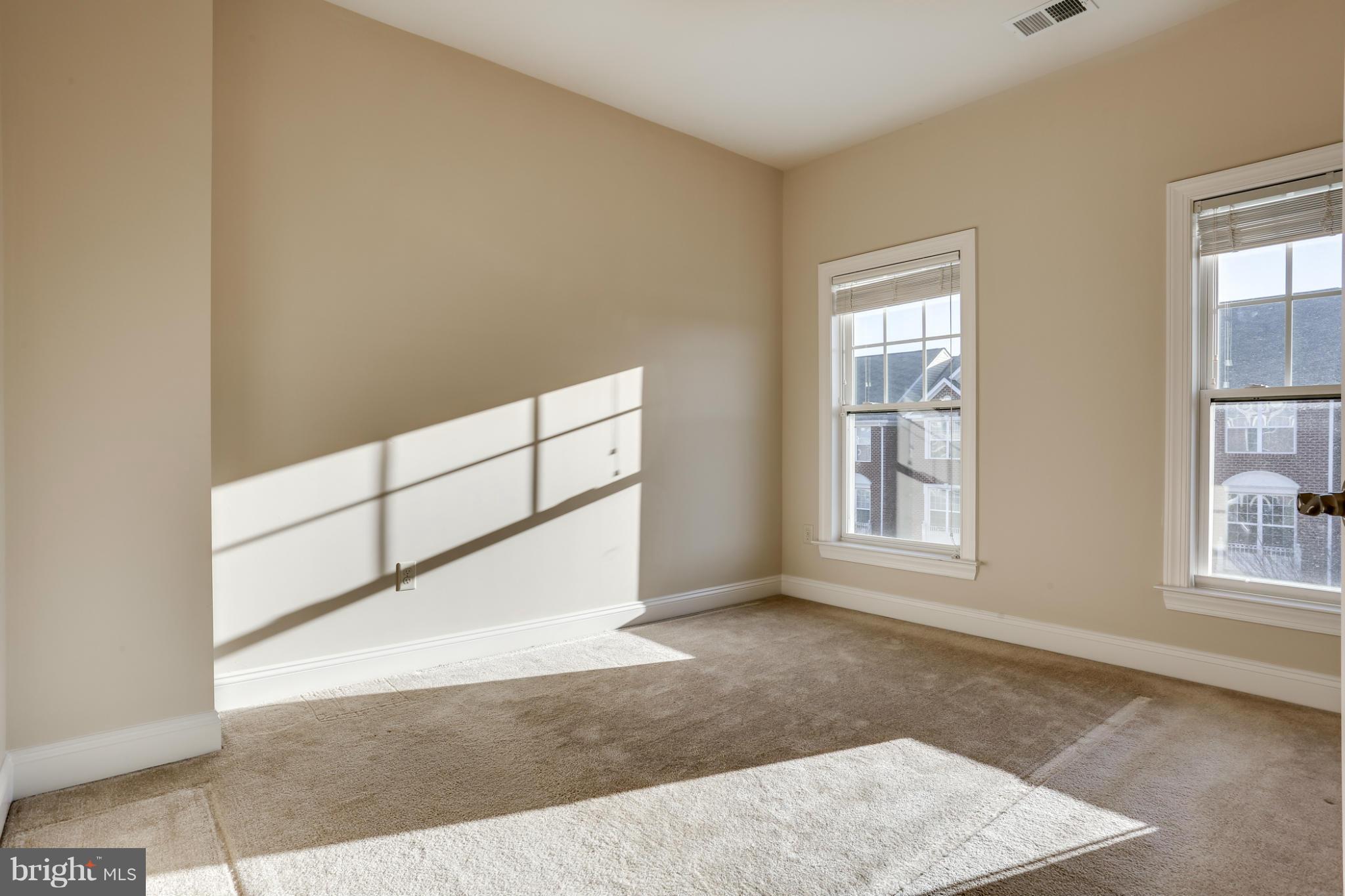 Empty carpeted room with two windows and strong sunlight casting shadows across the wall and floor.