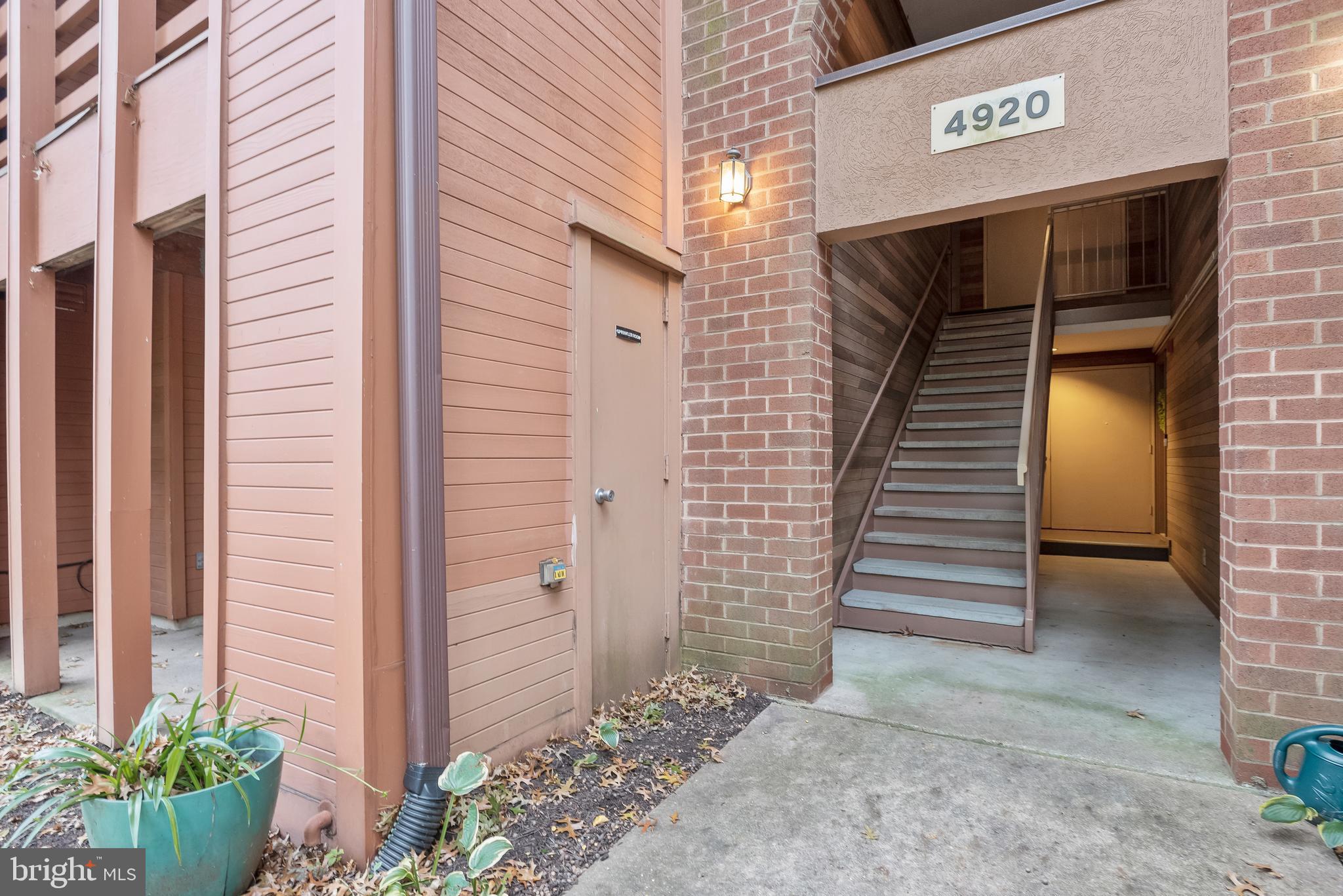 Property entrance with a covered staircase, brick columns, tan siding, and potted plants.