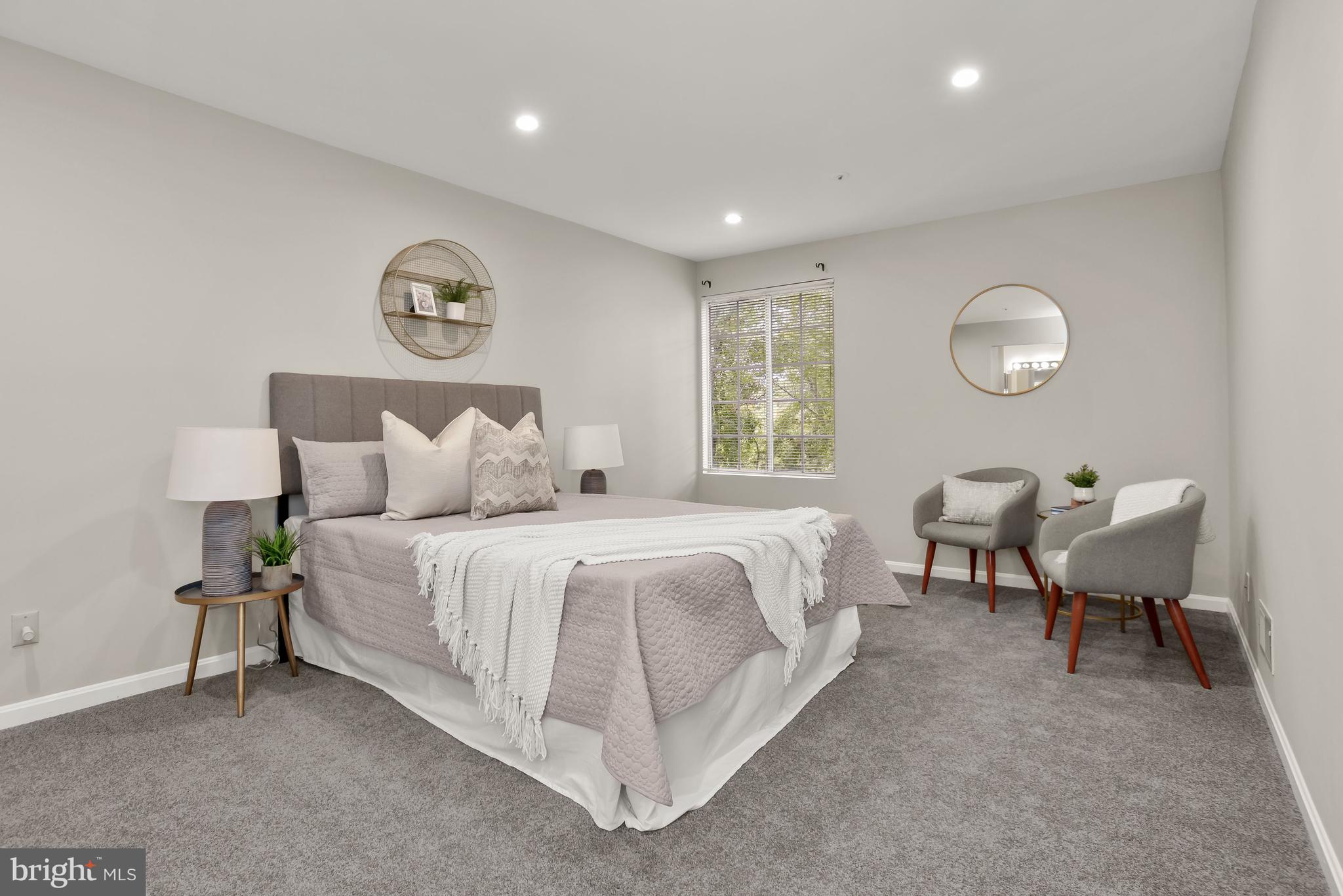 Staged bedroom with a gray-and-white bed and two chairs beside a window.
