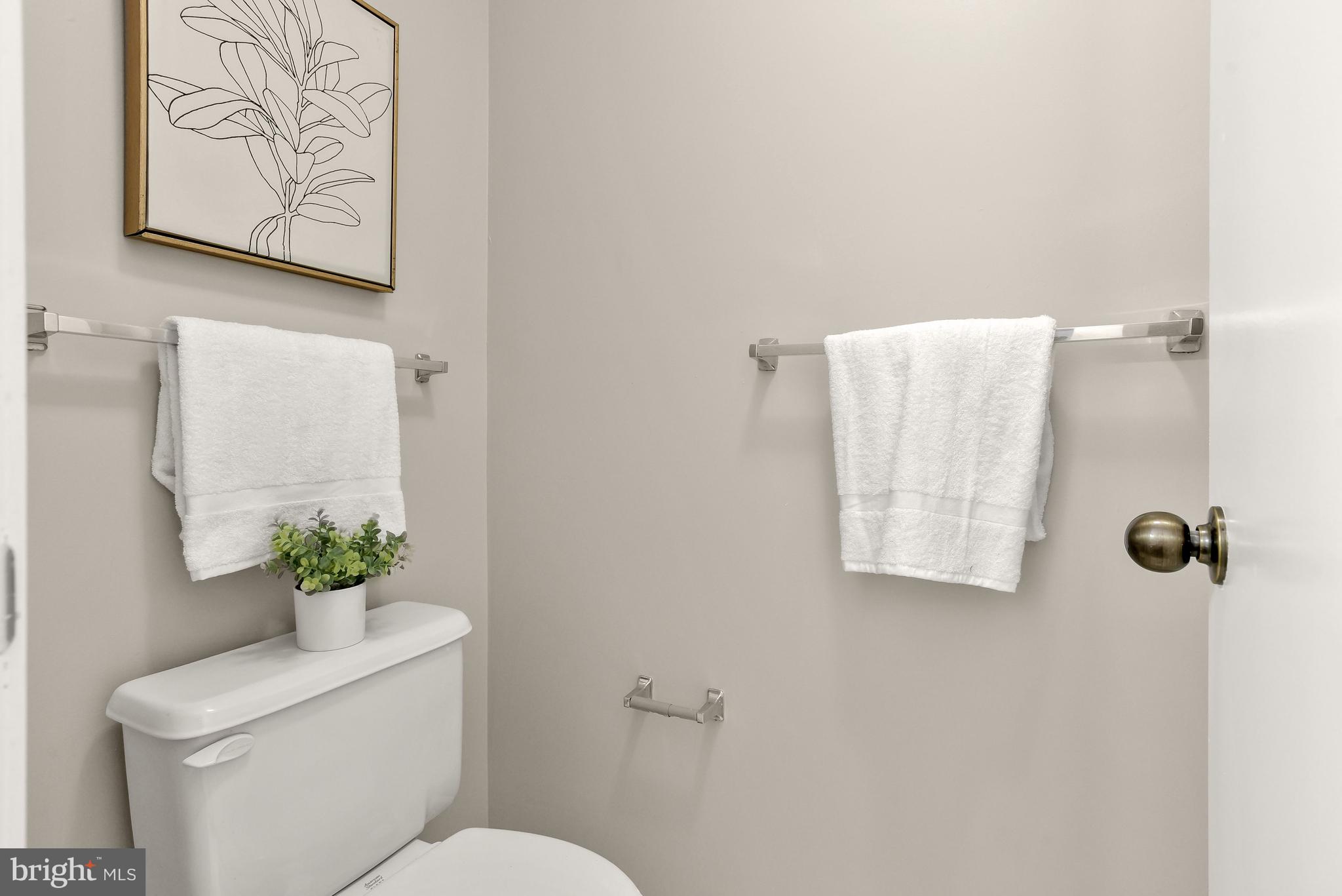 Staged bathroom with white towels, a toilet, framed botanical artwork, and a potted plant.