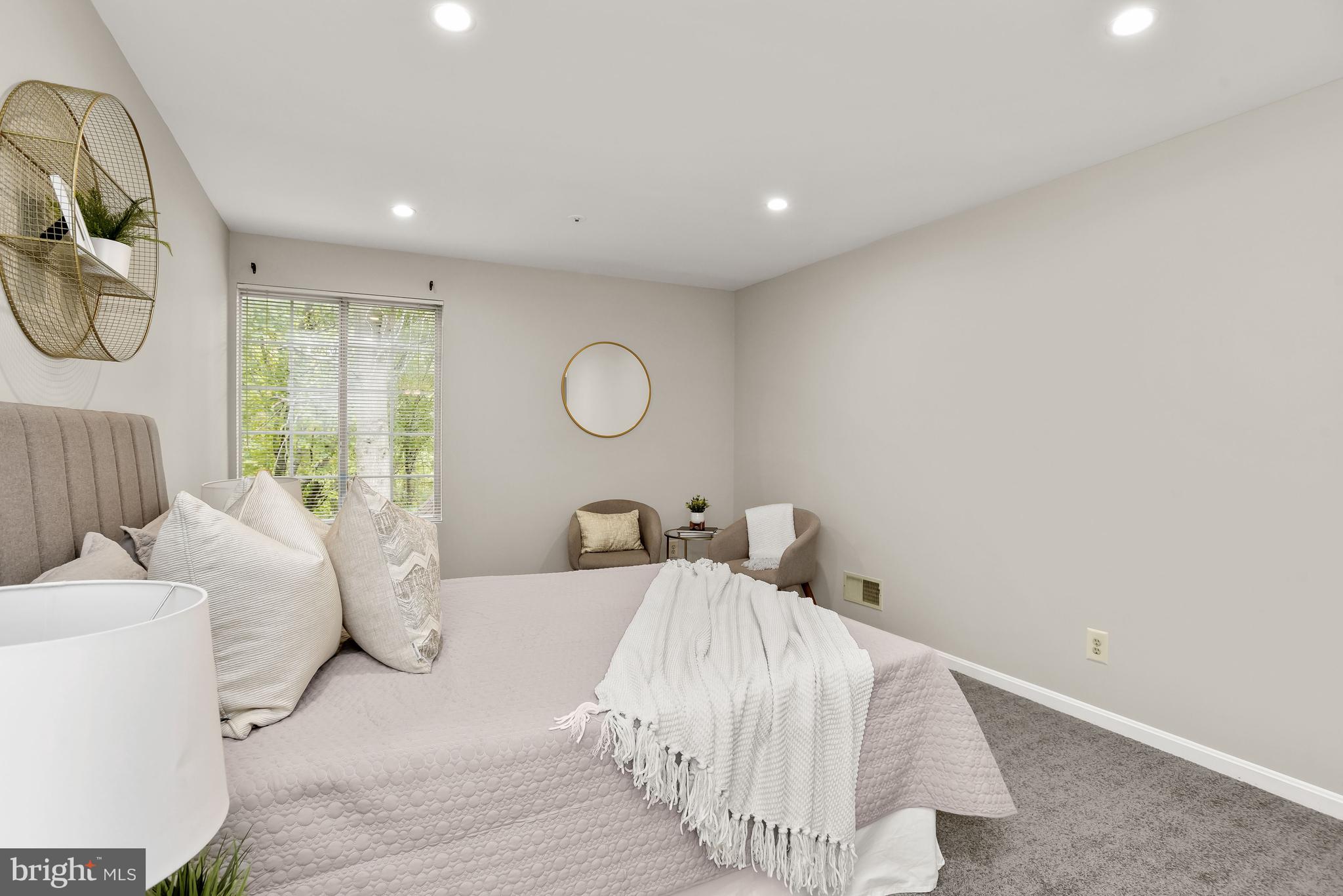 Furnished bedroom with a pale bed, white fringed throw, window blinds, and two upholstered chairs.