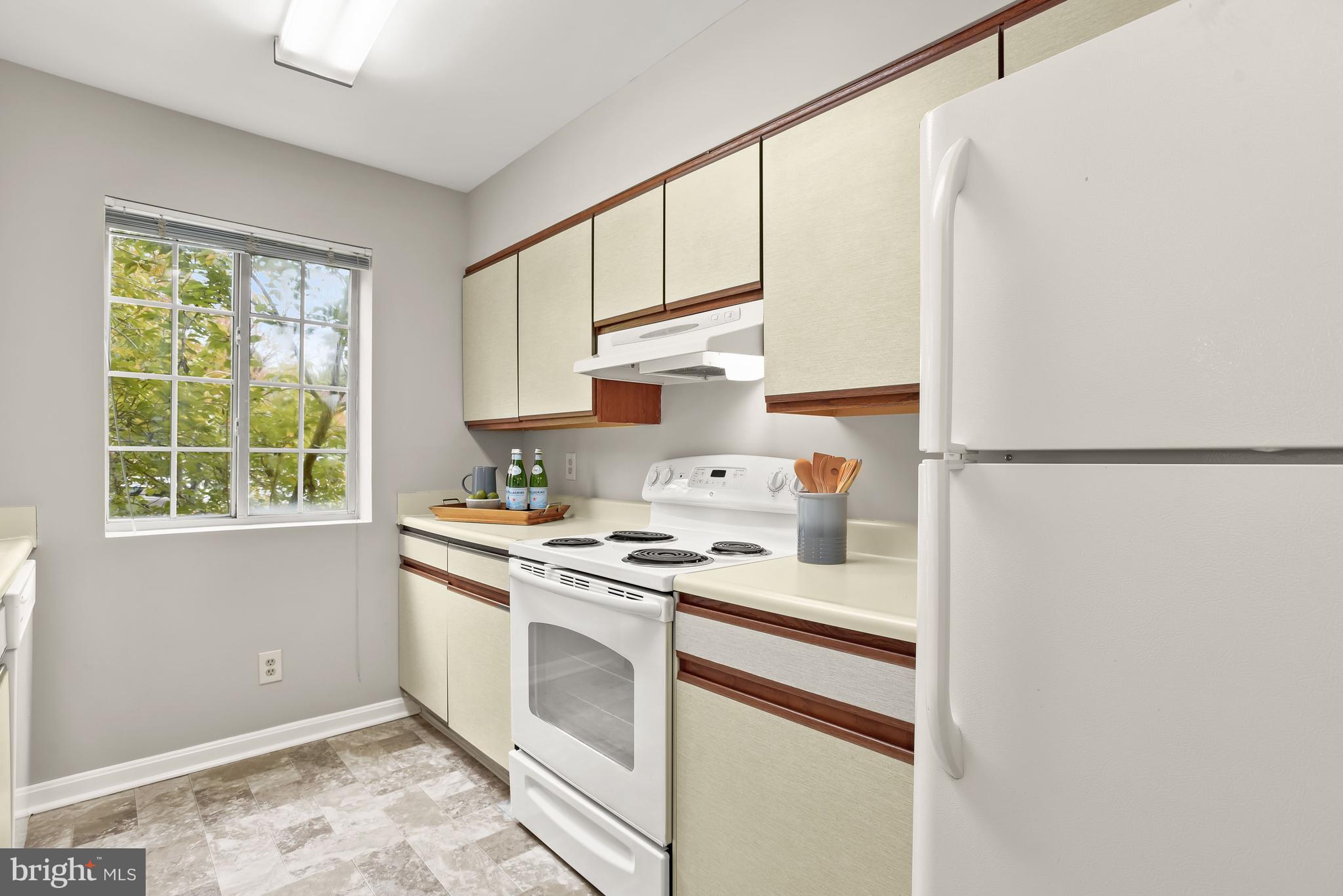 Kitchen with cream and wood-trimmed cabinets, white appliances, and a large window overlooking trees.