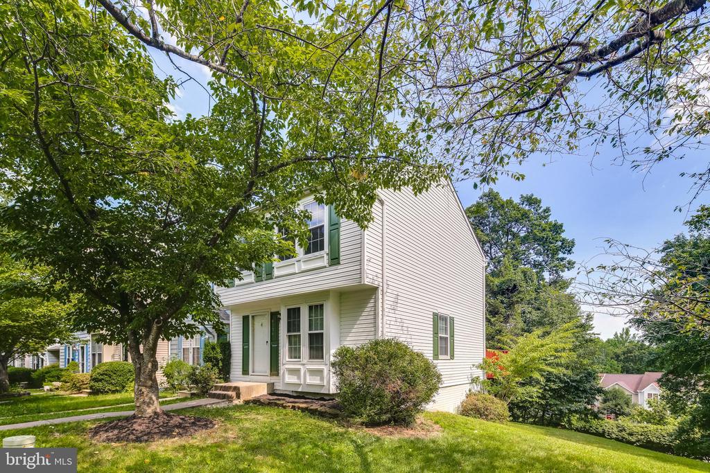 White two-story house with green shutters, framed by leafy trees on a sloping lawn.
