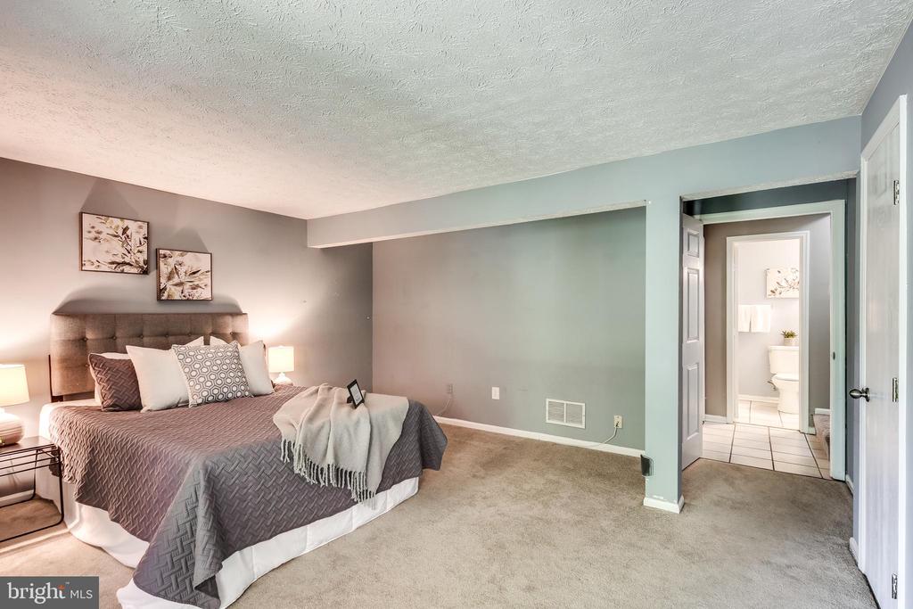 Staged bedroom with a large bed, gray carpet, and an adjoining bathroom visible through the doorway.