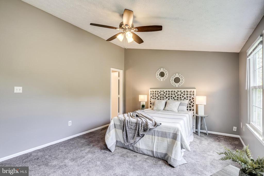Staged bedroom with a patterned white bed, gray carpet, ceiling fan, and large windows.