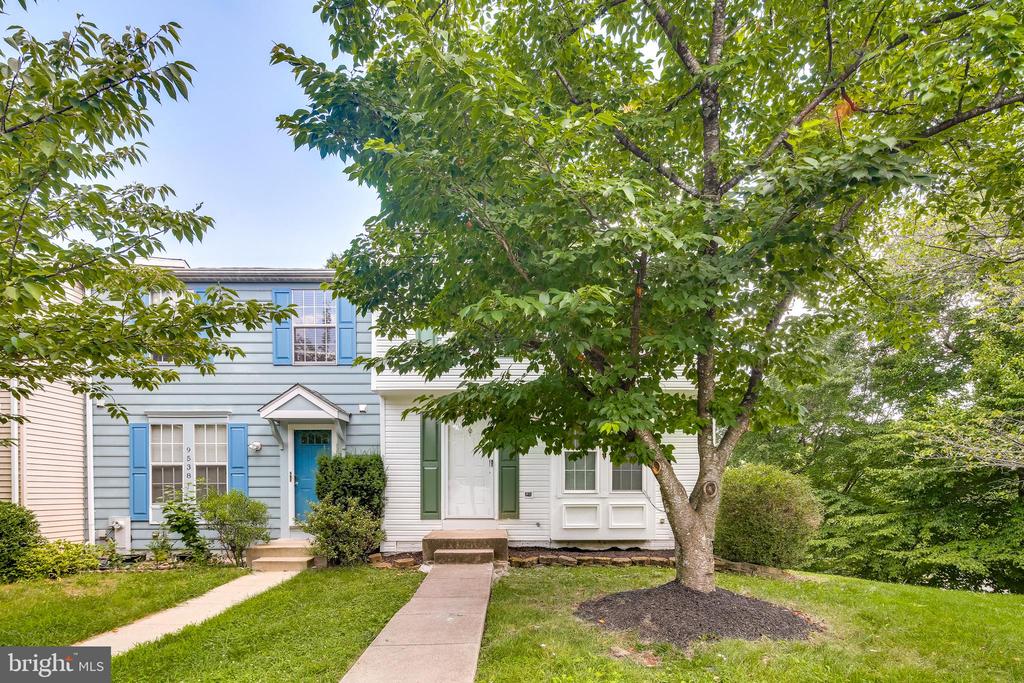 Two-story townhouse with blue shutters and entry door, shaded by a large tree.