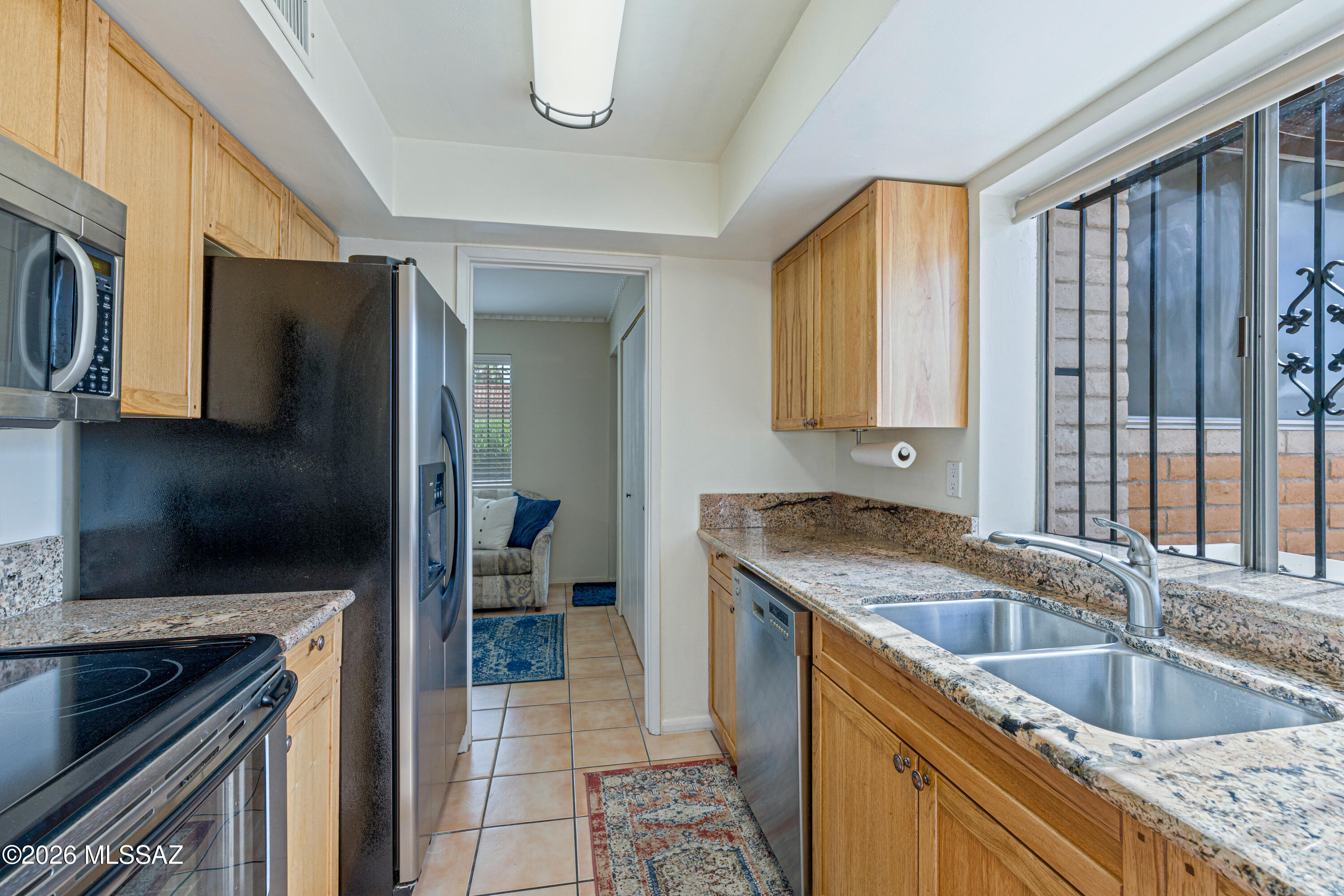 Kitchen with granite counters, light wood cabinets, and a double-basin sink beneath a barred window.