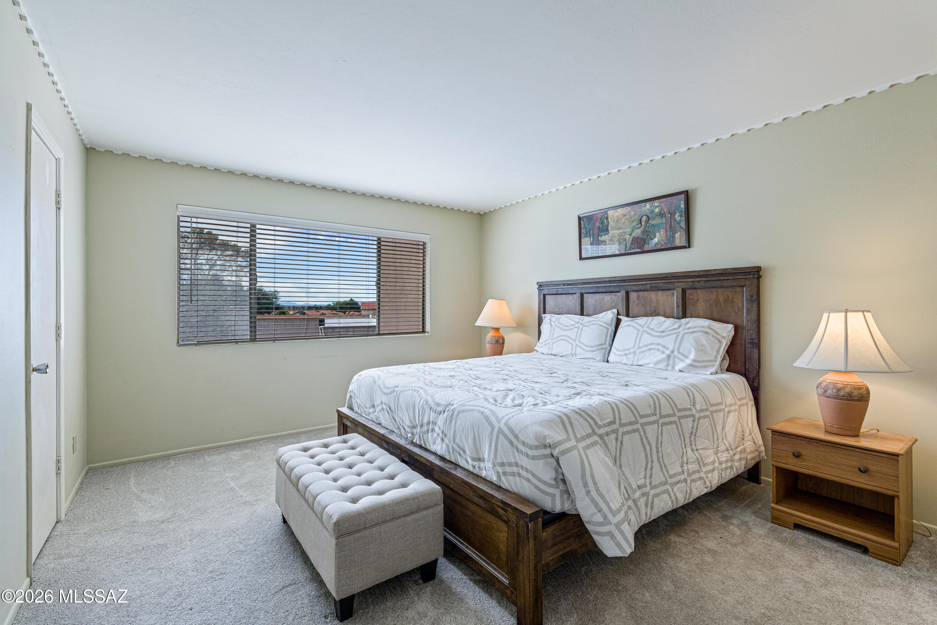 Bedroom with a dark wood bed, geometric bedding, tufted bench, and wide window with blinds.