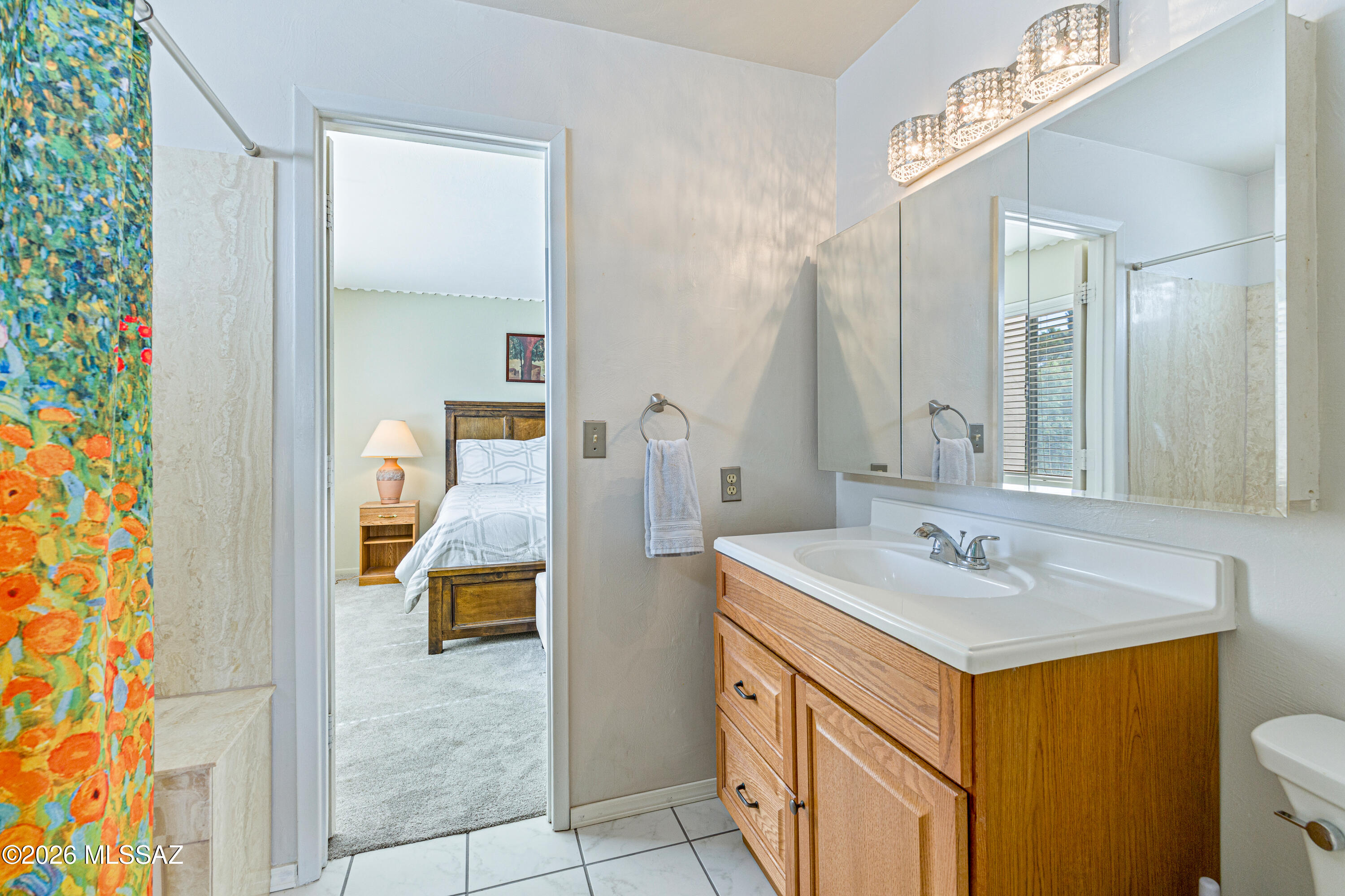 Bathroom with a wood vanity and colorful shower curtain; a furnished bedroom is visible through the doorway.